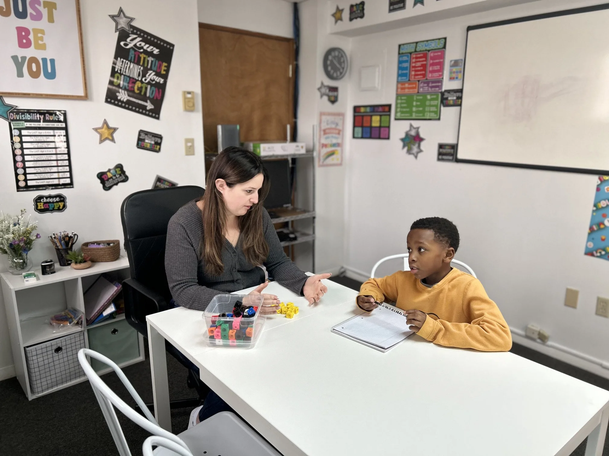 A woman and a young boy sitting at a white table in a classroom, engaged in a conversation. The classroom walls are decorated with colorful posters and educational materials, including a "Disability Rules" chart and various stars and motivational signs.