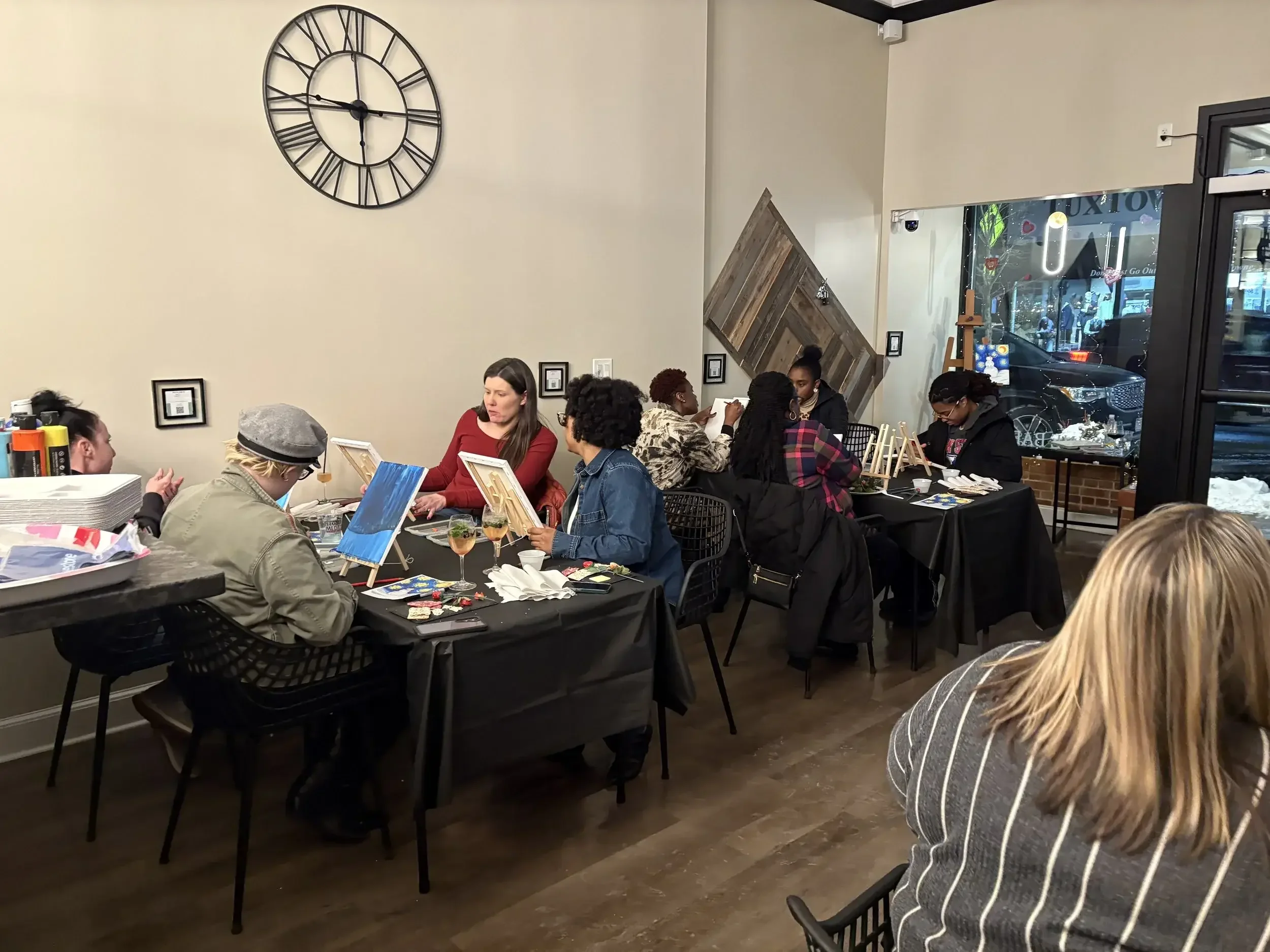 People sitting at tables inside a cafe or restaurant, engaging in conversation and painting on small canvases, with decorative items on the tables and a large clock on the wall.