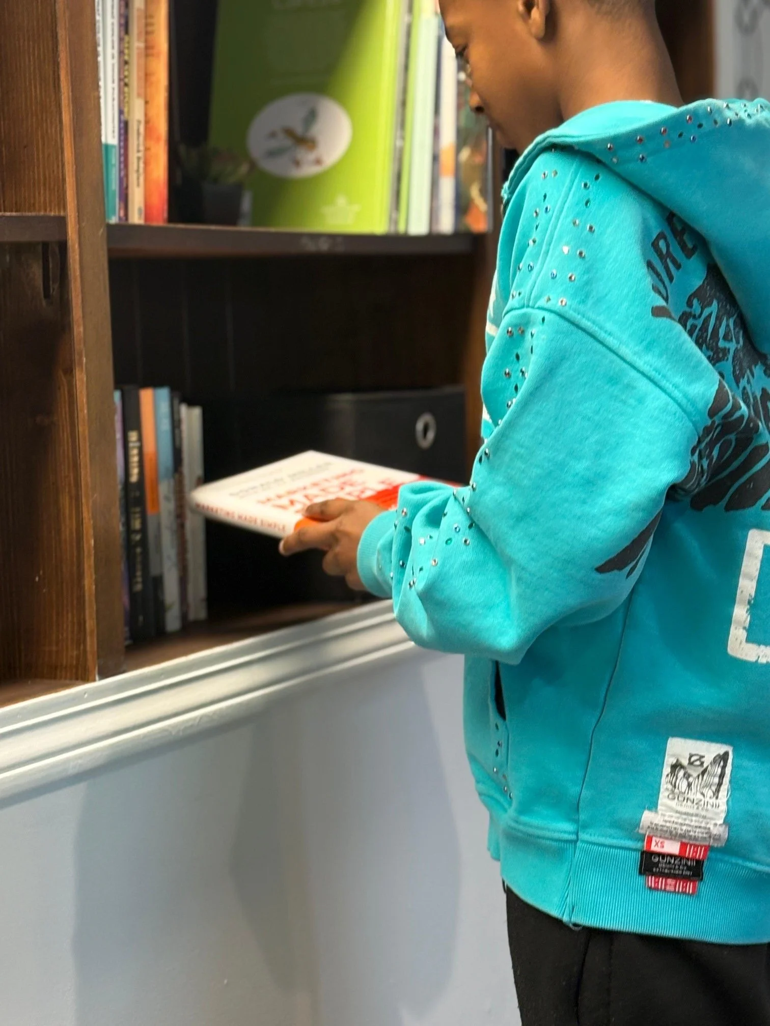 Teenager in teal hoodie reading a book at a library bookshelf.