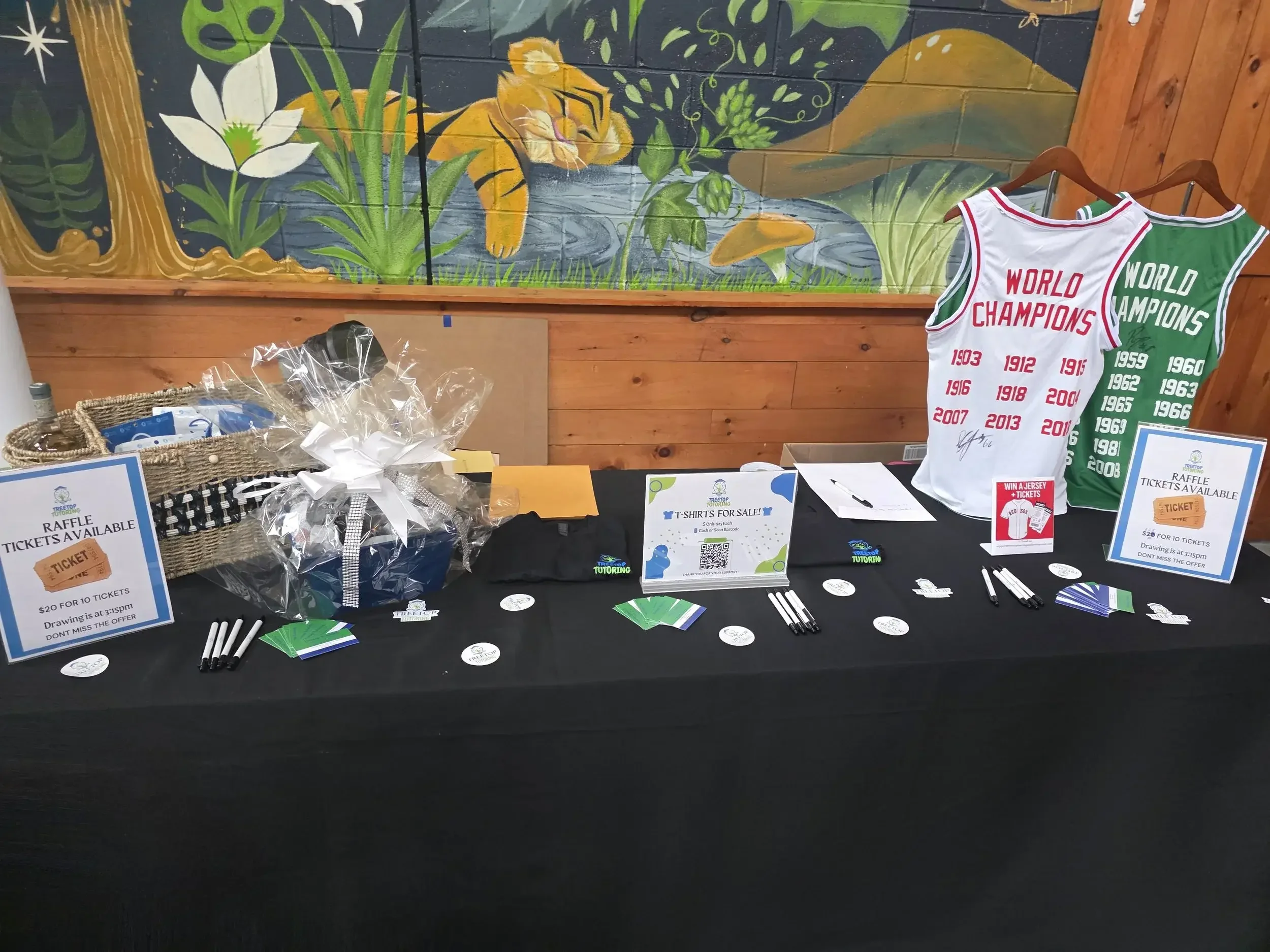 A colorful mural of a tiger resting by a pond, surrounded by green plants and mushrooms, painted on a brick wall. In front of the mural, there are raffle tickets, pens, and signs on a table, with sports jerseys hanging on a rack to the right.