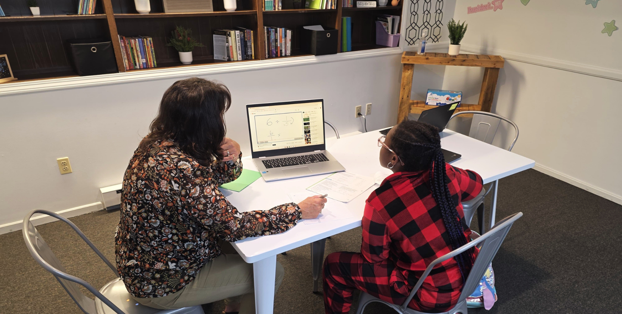 Teacher and student working together at a table in a classroom, with a laptop showing math problems and books on shelves in the background.