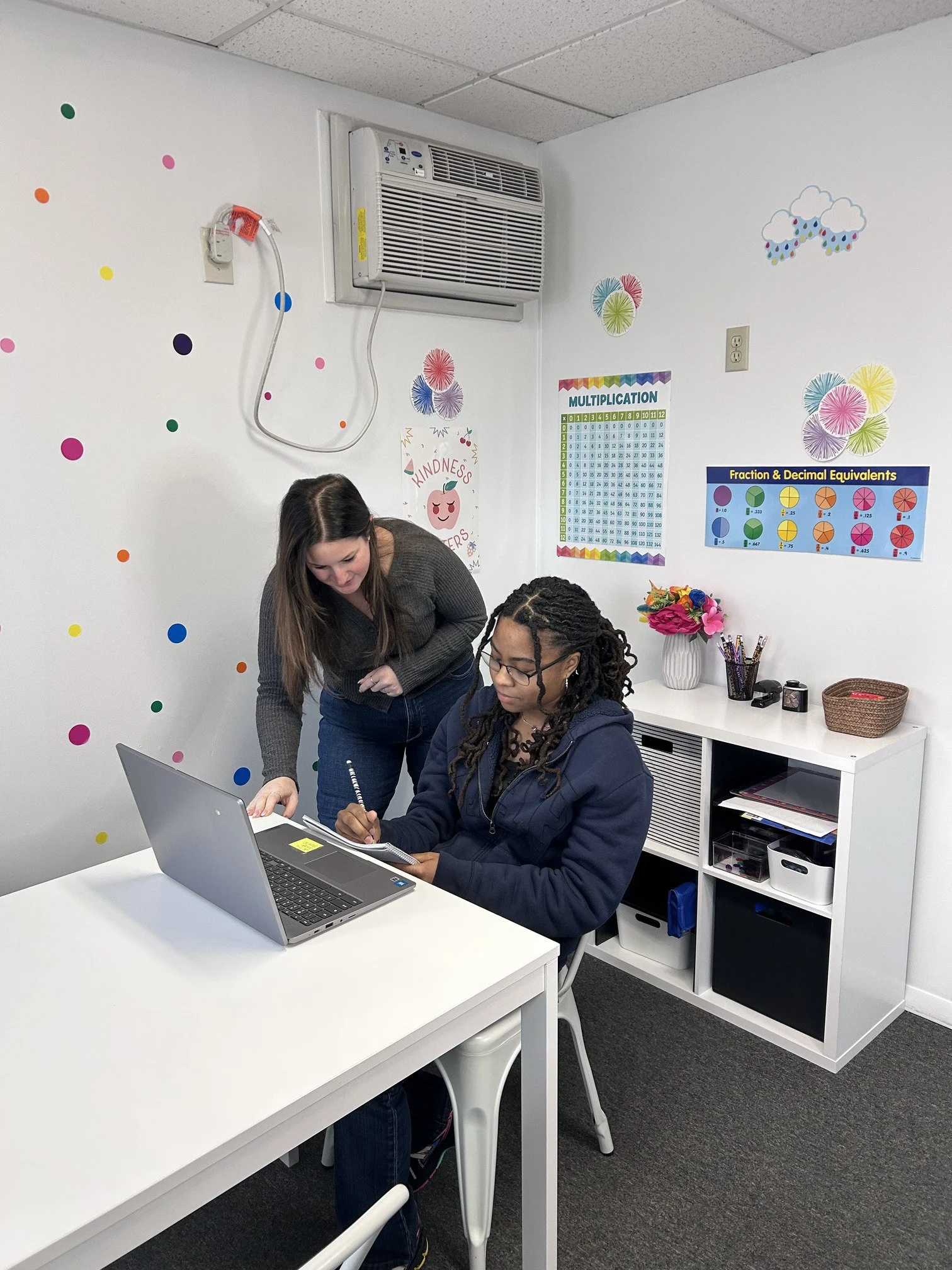 Two women working at a white desk in an office. One woman is sitting and looking at notes, the other woman is leaning over and pointing at the notepad. Wall with colorful stickers and educational posters about multiplication and fractions in the background, along with a small table holding a vase with flowers and some office supplies.