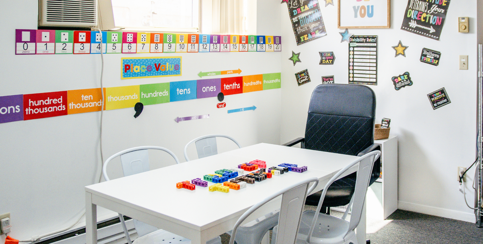 A classroom corner with educational posters and colorful number and place value charts on the wall, a white table with assorted colorful counting blocks, and a chair with a black cushioned backrest. There's a window with blinds and an air conditioning unit on the wall.
