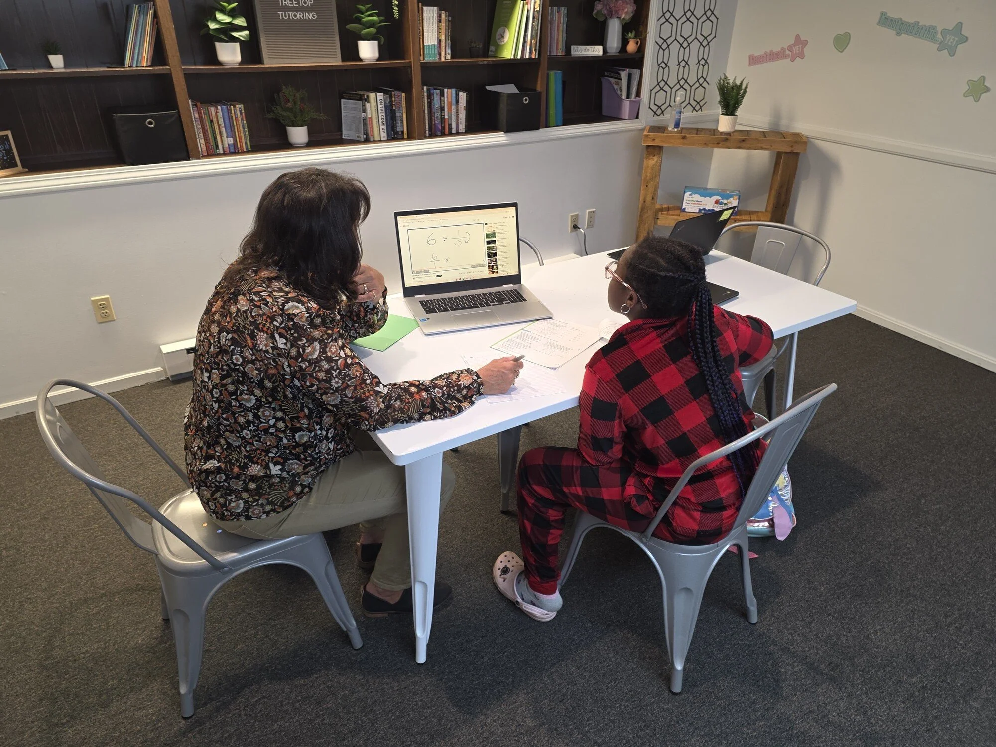 Two women sit at a white table in a room with a dark carpet. One woman with dark, curly hair wears a floral blouse; the other woman with braided hair wears glasses and a red and black plaid shirt. They are looking at laptops and papers. In the background, there is a bookshelf with books, potted plants, and decorative items, and the wall has colorful motivational stickers.