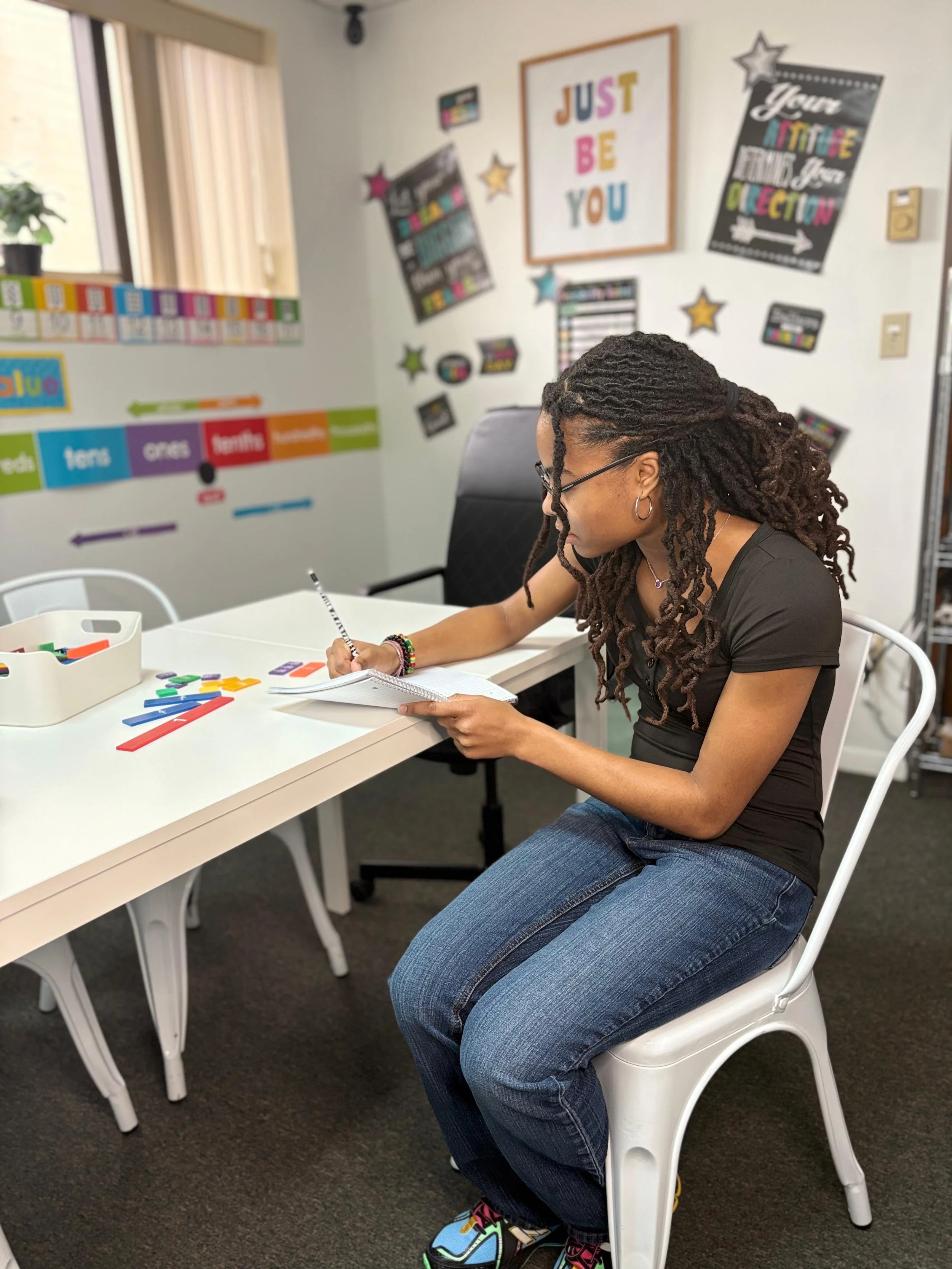 A person with long dreadlocks, glasses, and earrings sitting at a table in a classroom, writing in a notebook, with classroom decorations on the wall behind them.