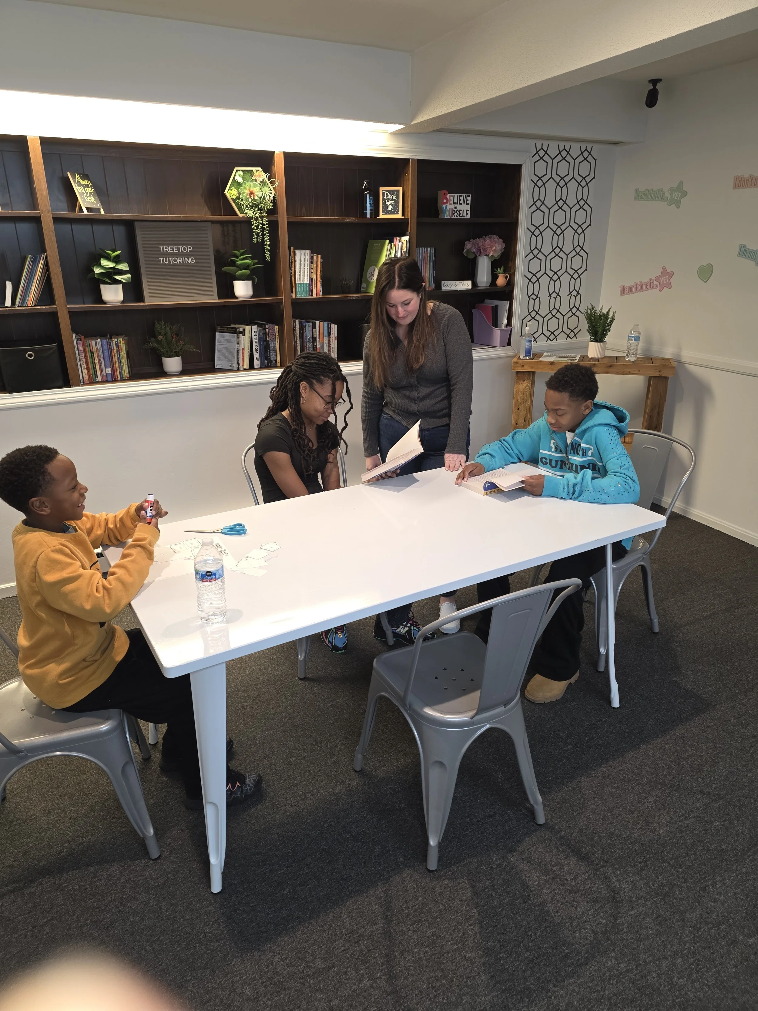 A classroom or study room with four children and an adult around a white table. Three children are sitting, engaged with books and papers, while an adult stands in the middle, looking at a book. The background features a bookshelf with books, potted plants, and decorative items, and walls with motivational quotes written in colorful text.