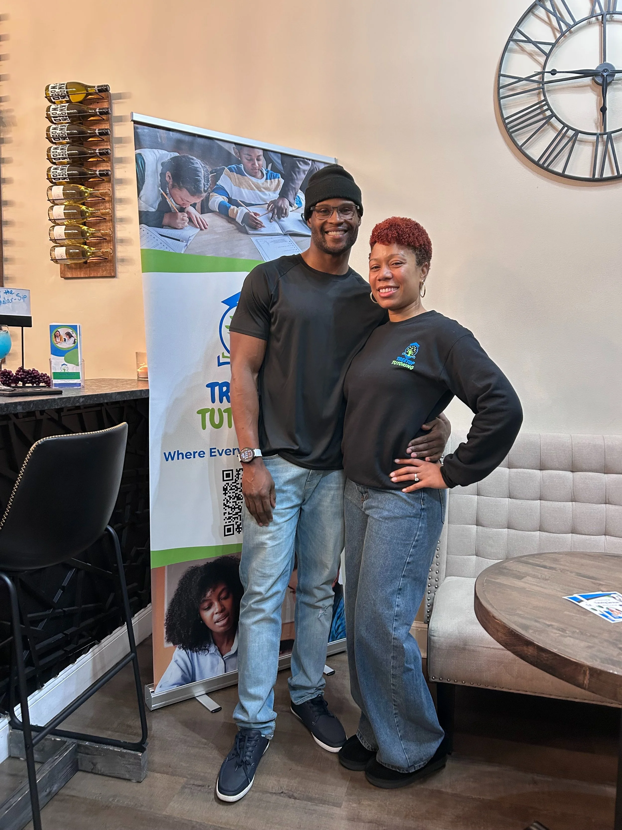 A man and a woman standing close together indoors, smiling at the camera, with a large clock on the wall behind them and a promotional banner to their left.