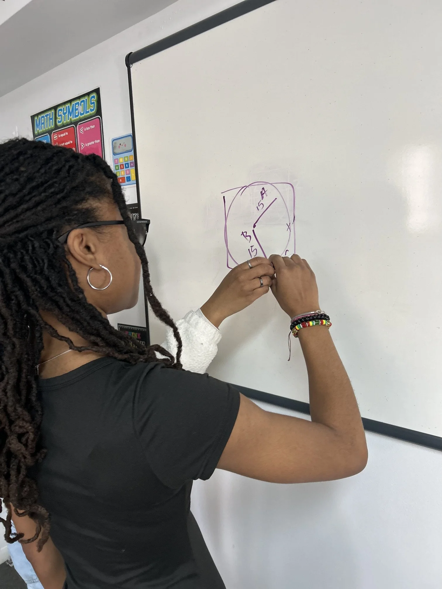 A person with long braided hair, glasses, and hoop earrings is placing a purple marker on a whiteboard, drawing a shape with angles and side lengths labeled.