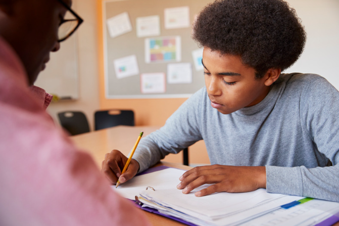 A young boy with curly hair studying at a table with a notebook and a pencil, while a person in a pink shirt looks on, in a classroom setting.