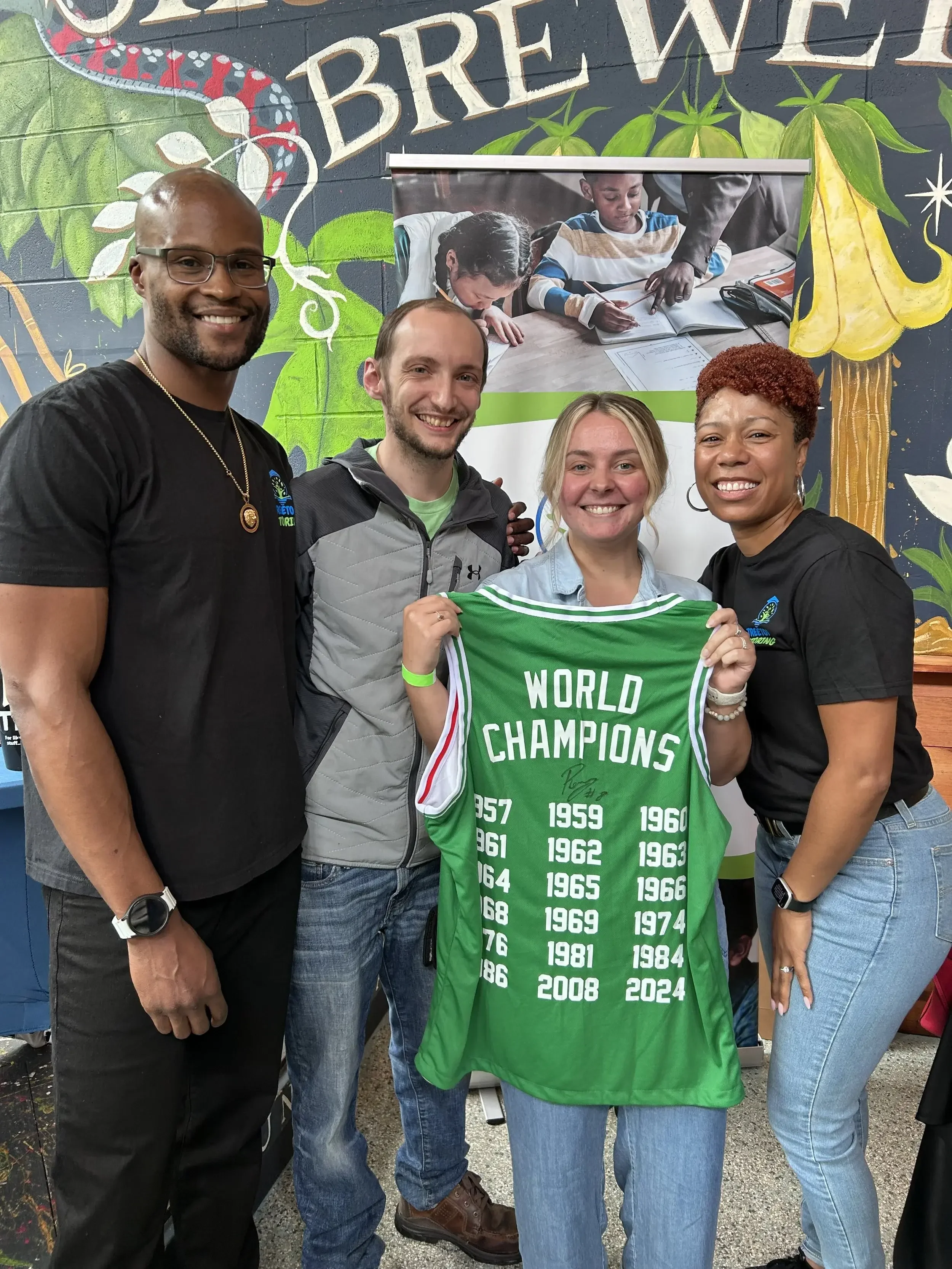 Four people standing together smiling, one woman holding a green sports jersey with years listed on it, in front of a decorative background and banner featuring children studying at a desk.