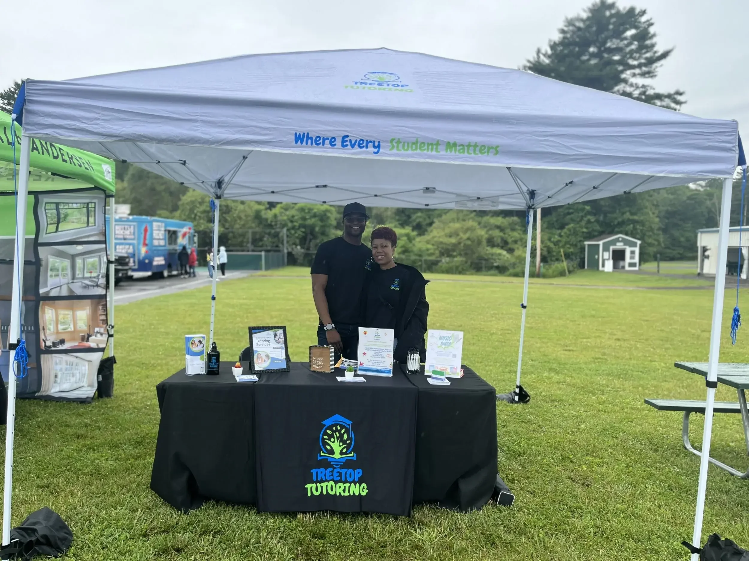 An outdoor event with a Treetop Tutoring booth set up under a white canopy. Two smiling people stand behind a table displaying pamphlets and small items. The canopy has the Treetop Tutoring logo and the slogan "Where Every Student Matters." In the background, there are trees, a grassy field, and other booths.