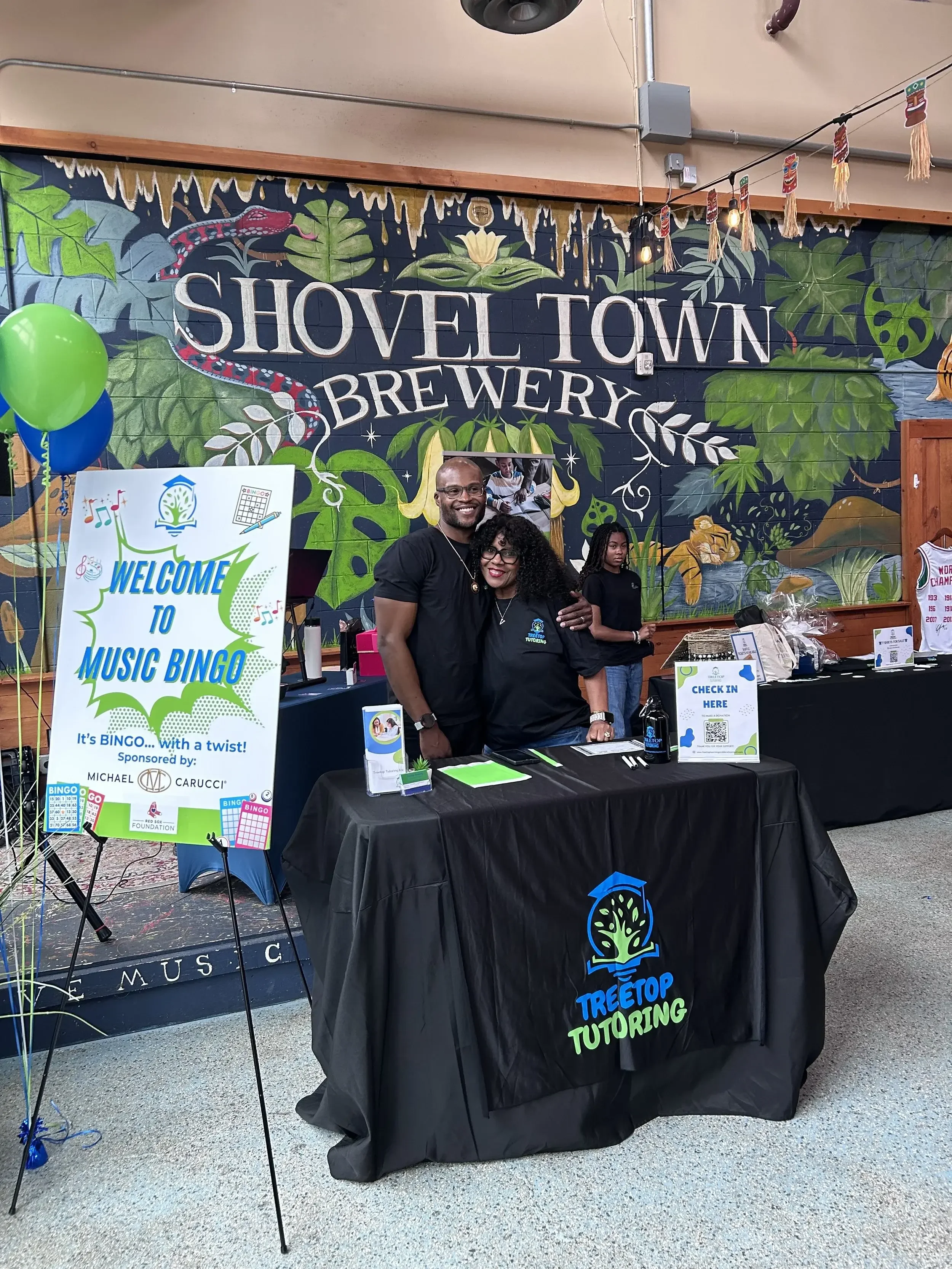 A booth for Treetop Tutoring at an indoor event with a mural background reading "Shovel Town Brewery." The booth has a black tablecloth with the tutoring company's logo, some promotional materials, and a sign-in sheet. Two smiling people are standing behind the table, while a woman stands further back. Balloons and decorative banners are also present.