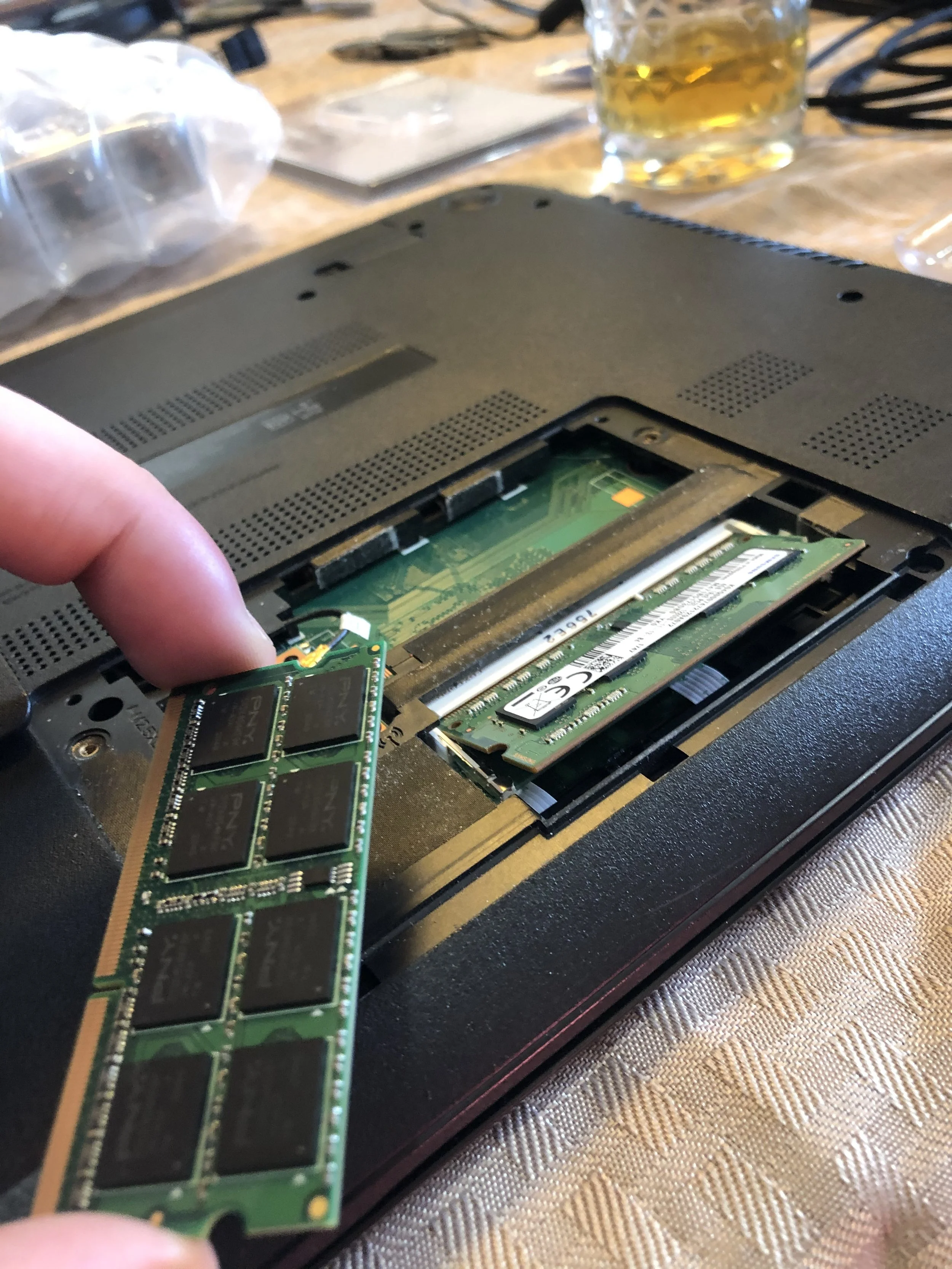 Person installing a RAM stick into a laptop's memory slot on a wooden table with drinks in the background.
