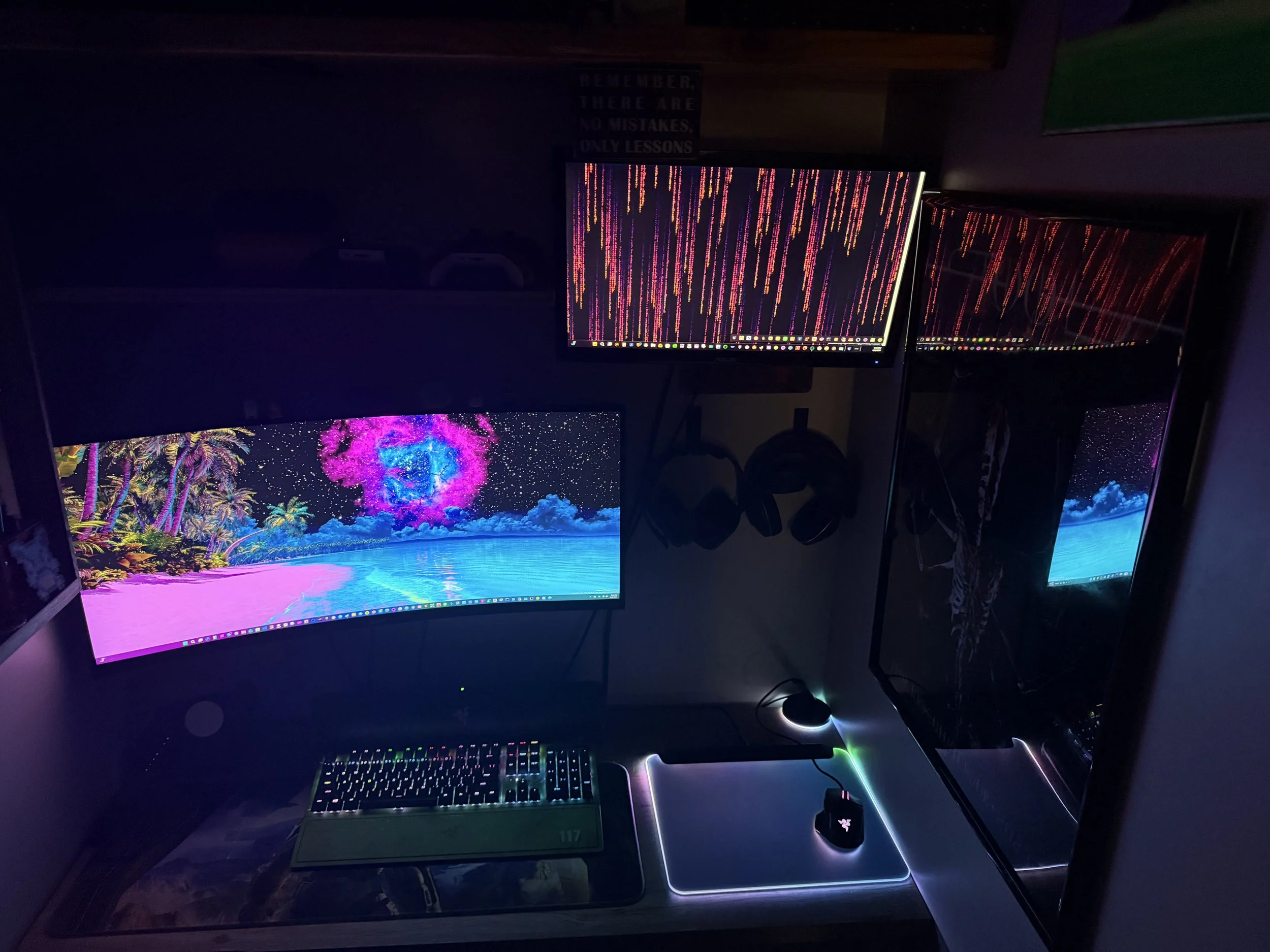 A gaming setup with three monitors displaying colorful, abstract images, a black keyboard with RGB lighting, a mouse on a glowing mouse pad, and headphones hanging on the wall.