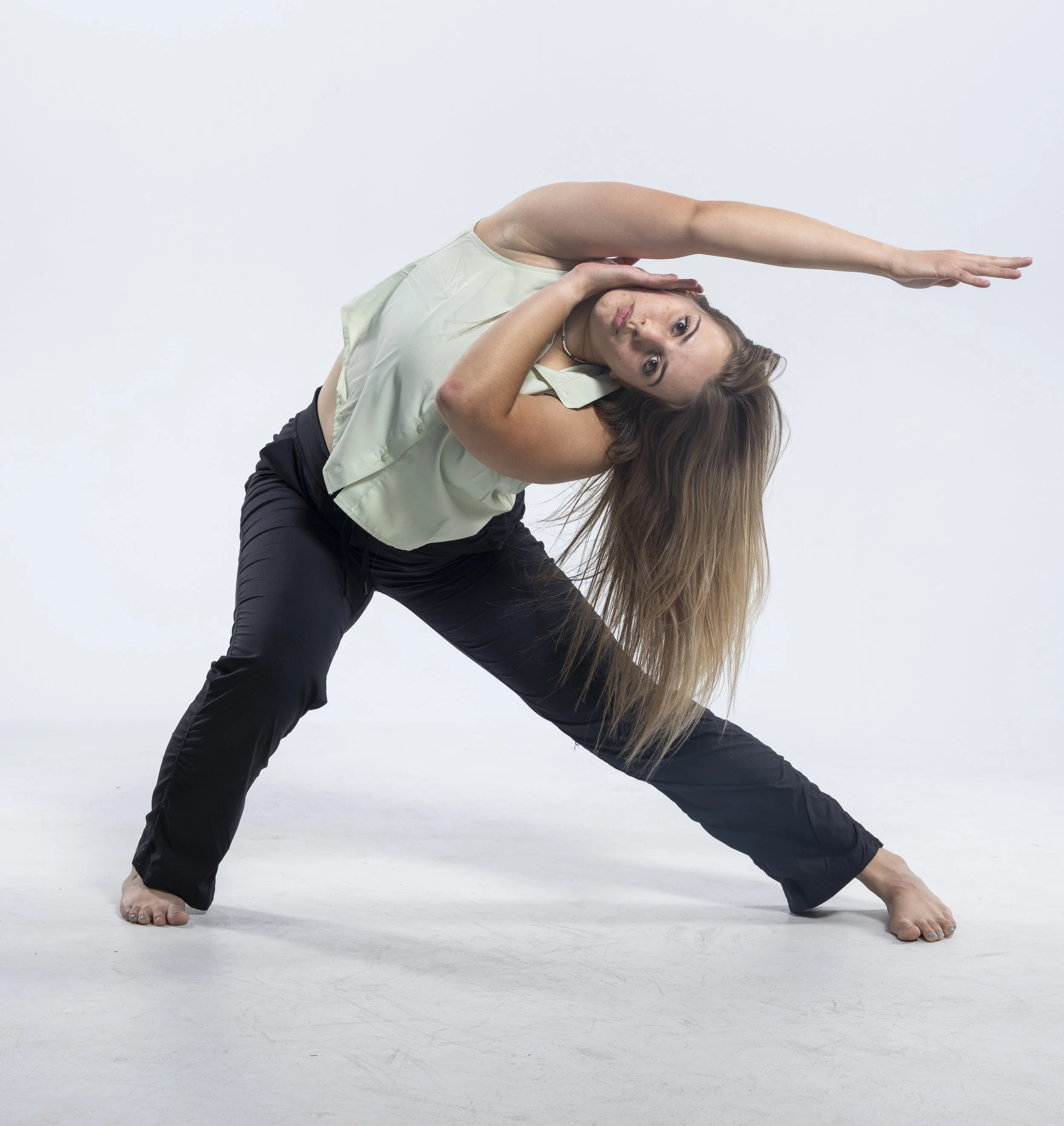 A woman in a green tank top and black pants performing a yoga pose on a white floor, with her body bent to the side, stretching her arm overhead and her other arm resting on her neck.