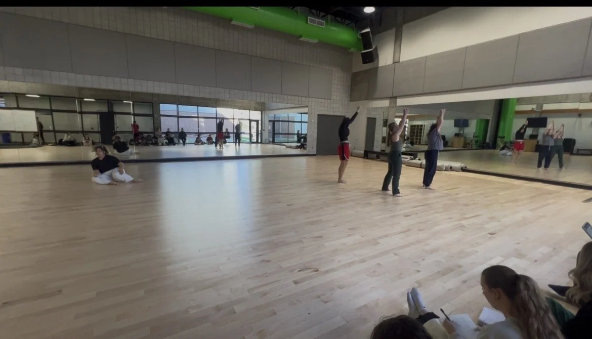 A dance class in a studio with wooden floors, a large mirror, and students practicing dance moves while others sit at the side and observe.