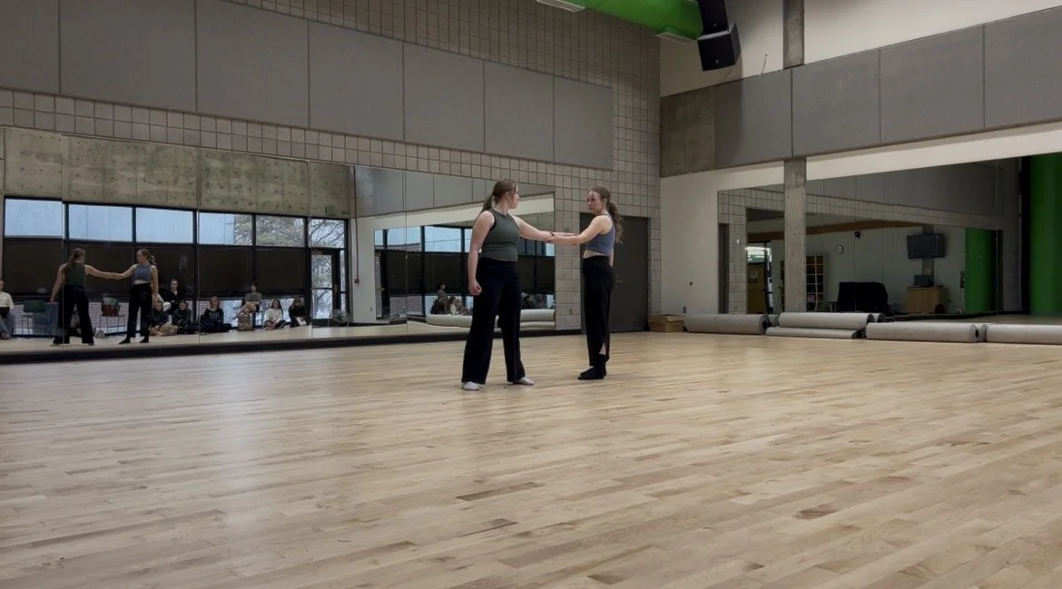 Two women holding hands and dancing in a dance studio with large mirrors, wooden floor, and other people sitting and watching in the background.