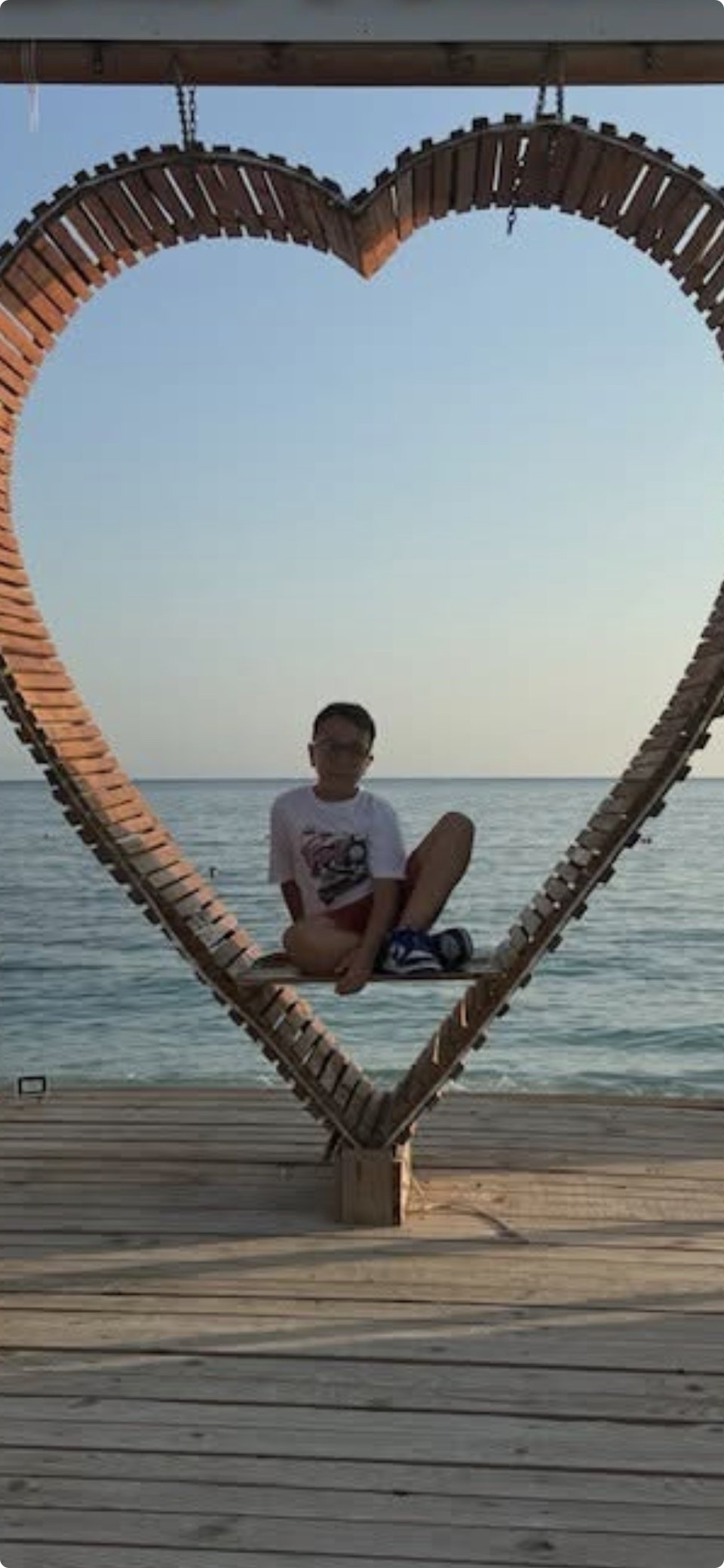 A young boy sitting on a bench shaped like a heart, overlooking the ocean at sunset.