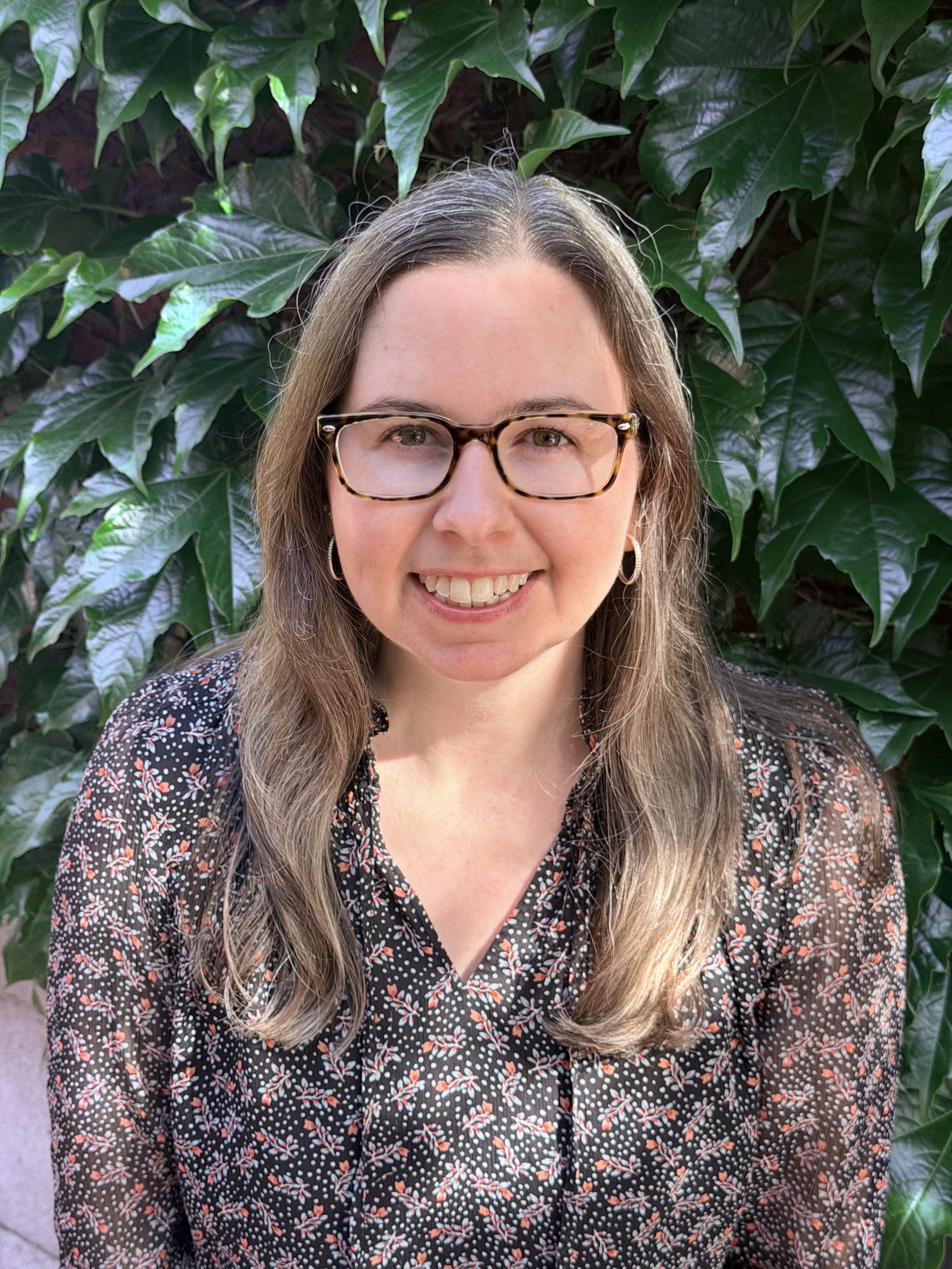 A woman with long wavy hair and glasses smiling in front of green leafy plants.