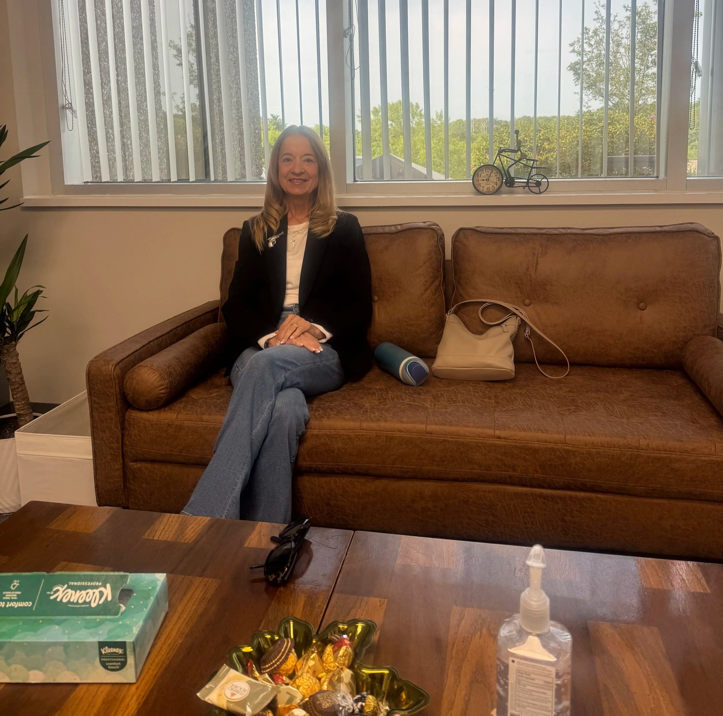 Woman sitting on a brown leather couch in front of a large window with vertical blinds, with trees and greenery visible outside. On the table in front of her are a box of Kleenex tissues, a bag of chocolates, a bottle of hand sanitizer, and some other items.