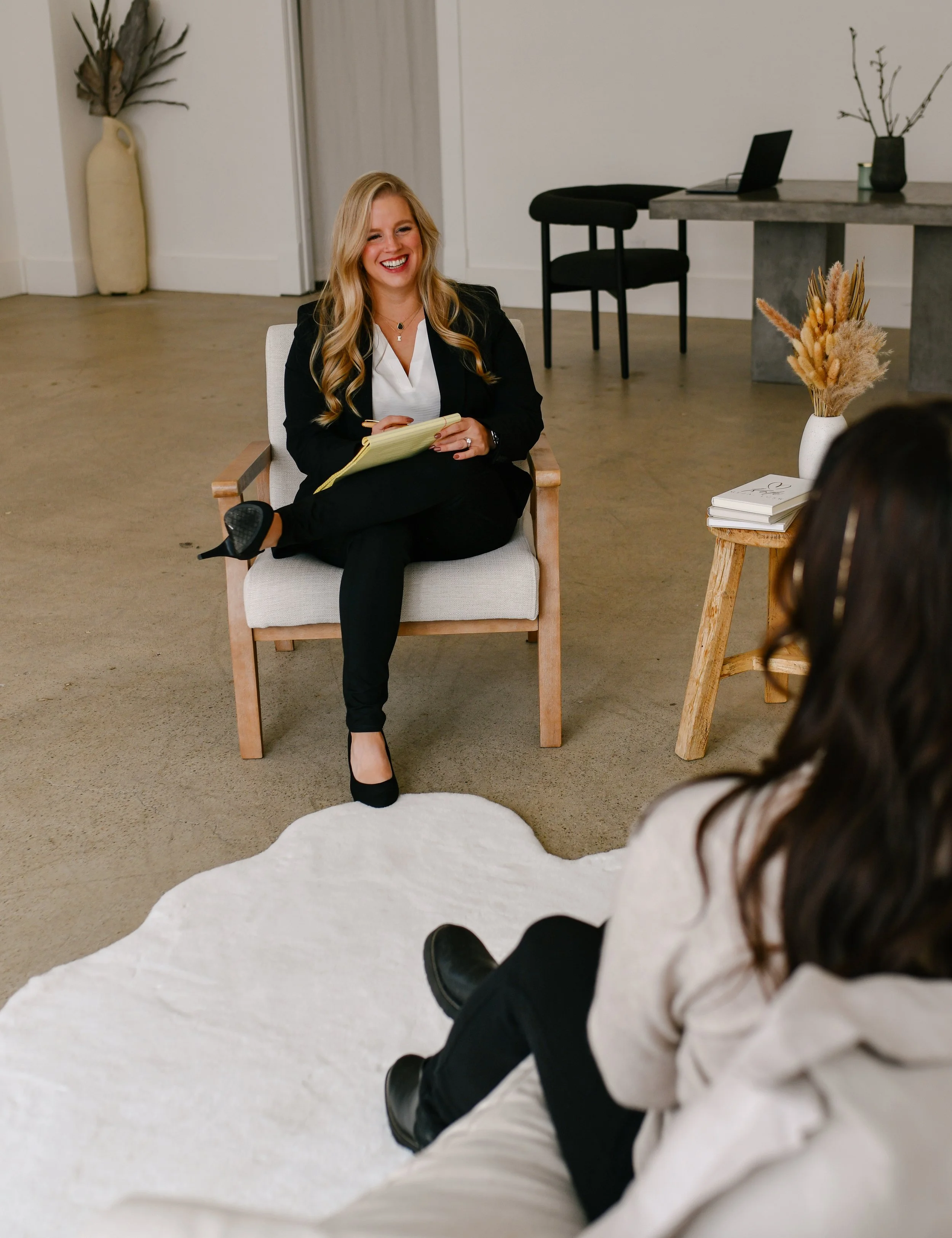 A woman with blonde hair is sitting on a chair, smiling, holding a notebook, appearing to be in conversation with someone out of frame. The setting is a minimalistic room with a white wall, decorative vases, and a table with a laptop and books.