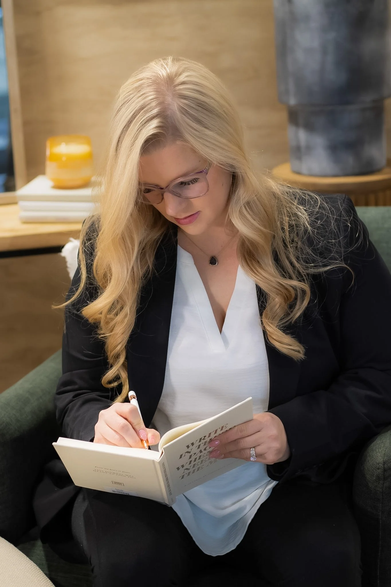 A woman with long blonde hair and glasses is sitting on a green armchair, writing in a book with a pen. She is dressed in a white top and black blazer, with a ring on her left hand. Behind her is a wooden table with a yellow bowl and some stacked books, and a decorative stone sculpture in the background.