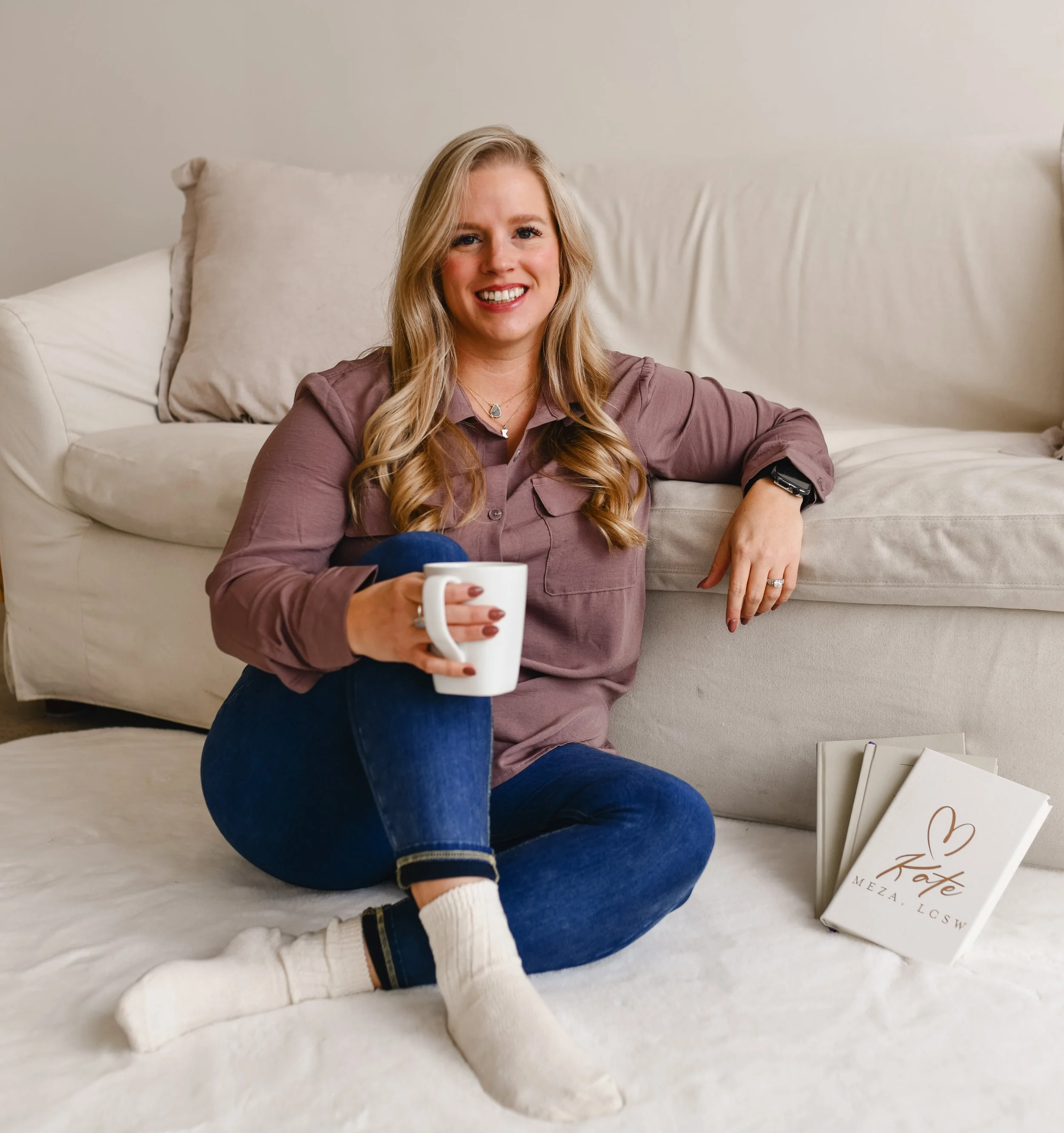 Woman with long blonde hair sitting on a white couch, holding a white mug, smiling at the camera, with books and papers on the floor nearby.