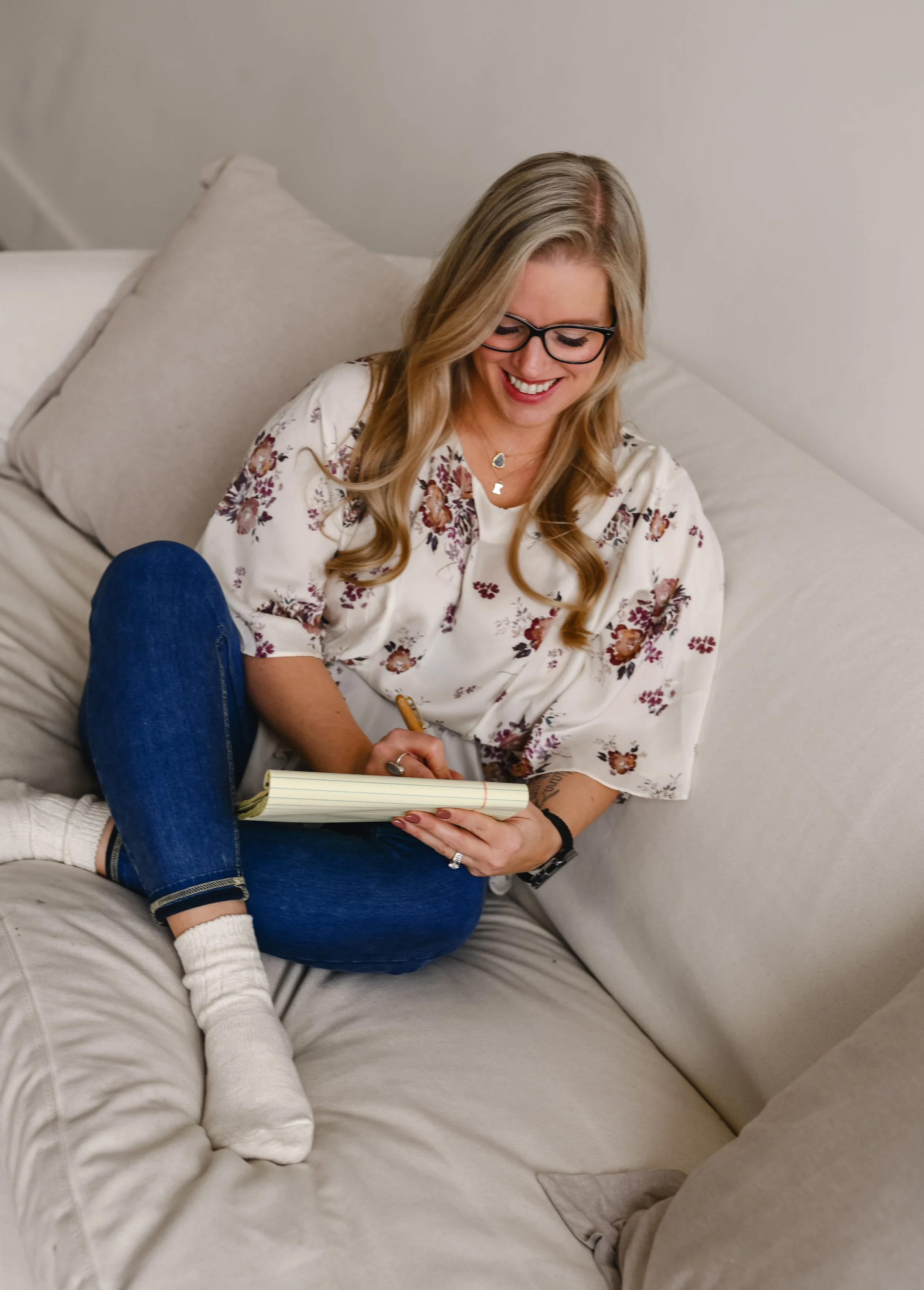 A woman with blonde hair, glasses, and a floral top is sitting on a beige couch, smiling while writing in a notebook. She has a bracelet on her wrist and a necklace, and is wearing blue jeans and white socks.
