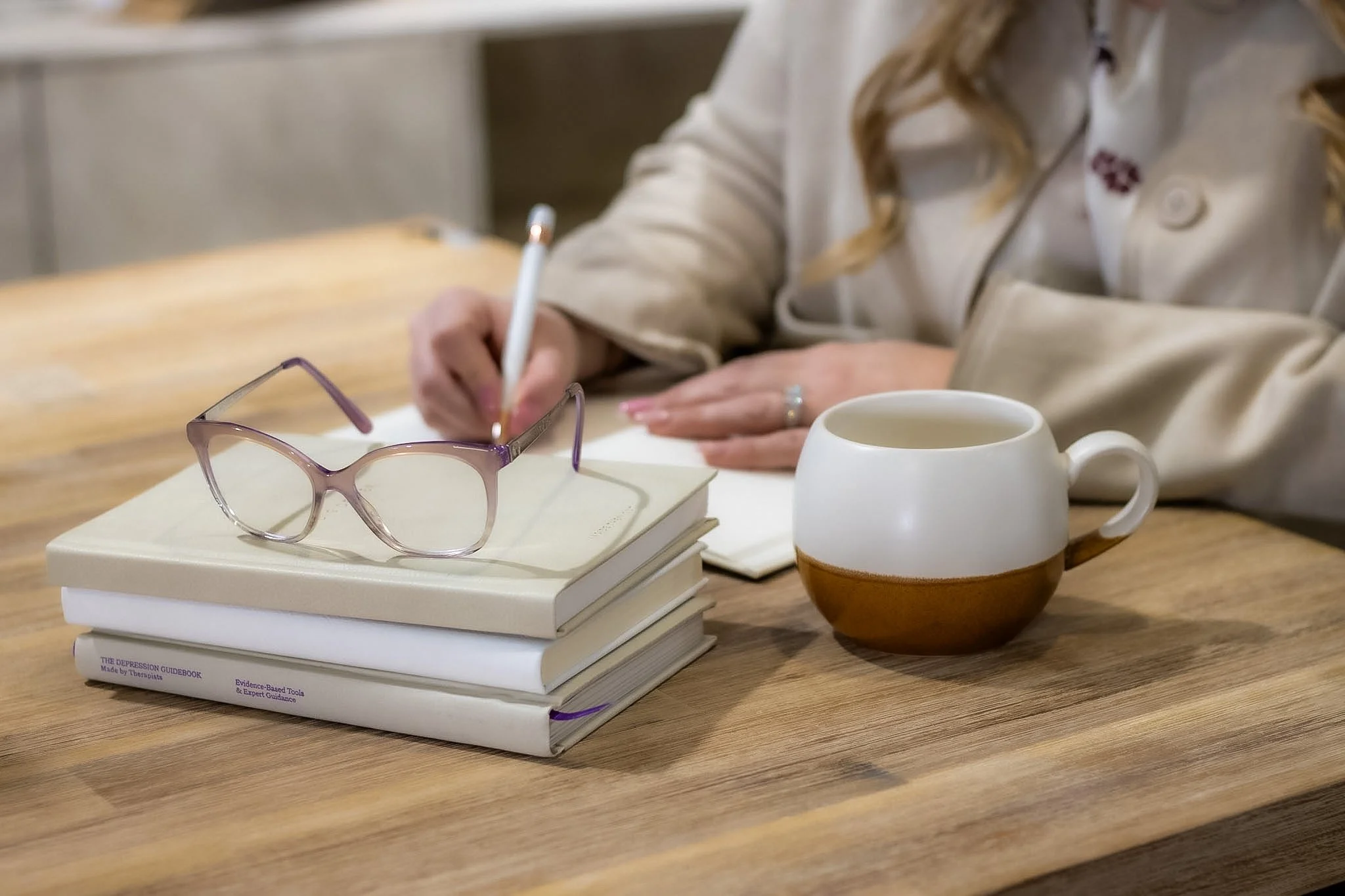 A stack of four books on a wooden table, with purple-framed eyeglasses on top, a white mug nearby, and a person in a beige jacket writing in a notebook.