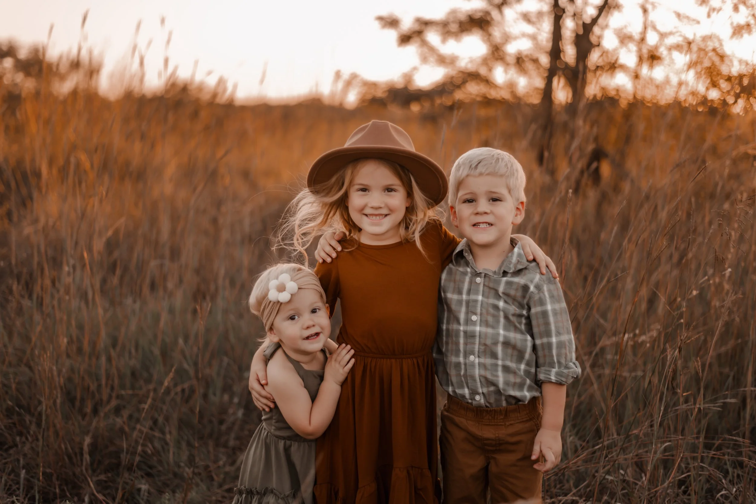 Three children standing in a field with tall grass at sunset, smiling and hugging each other. The middle girl wears a brown dress and a wide-brimmed hat, the boy on her right wears a plaid shirt, and the youngest girl on her left wears a green dress with a white flower headband.