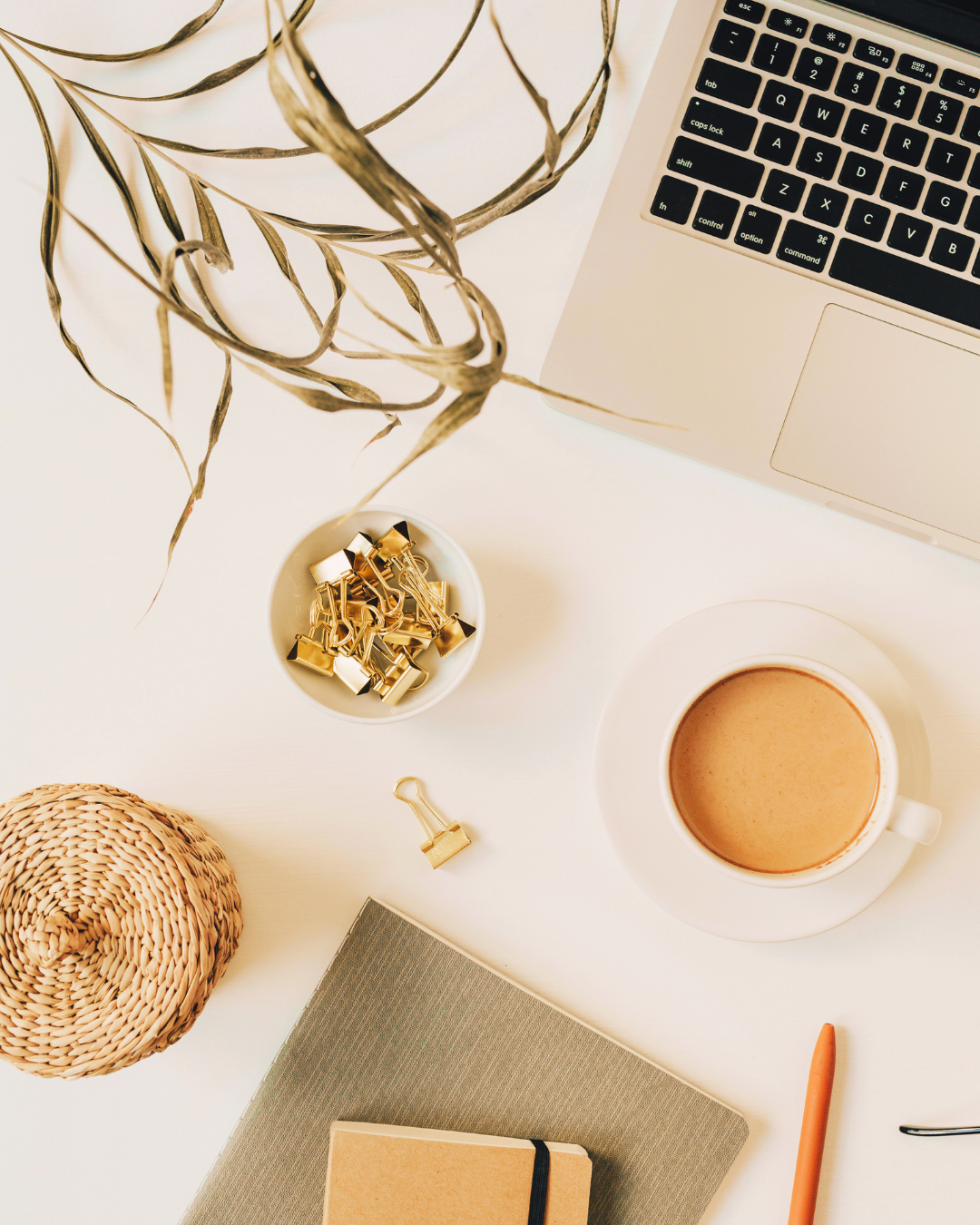 Top-down view of a workspace with a laptop, a cup of coffee, a notebook, a pen, gold binder clips in a bowl, a dried plant, and a woven coaster on a white desk.