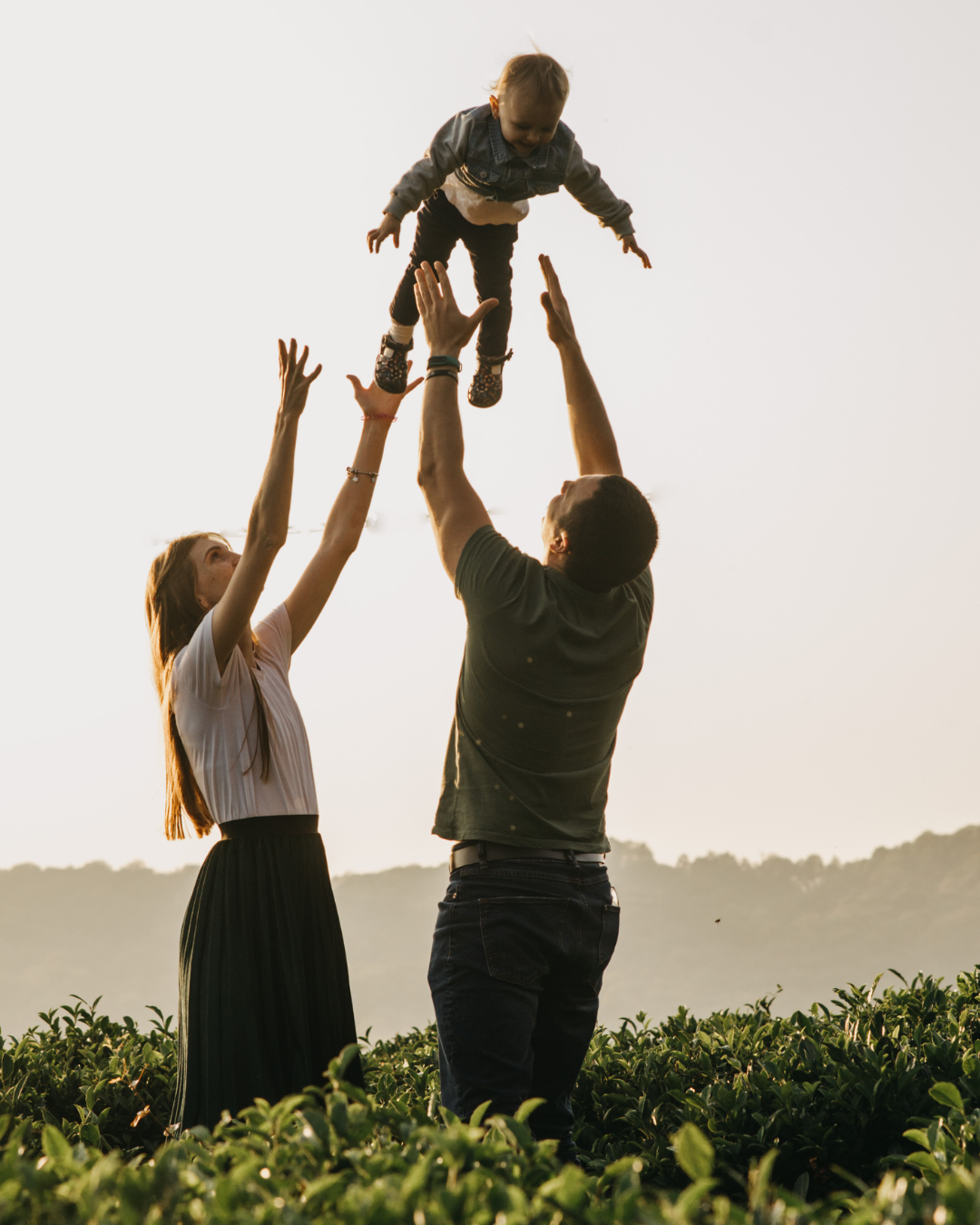 A family of three outdoors in a field during sunset, with the father throwing a child into the air while the mother watches and reaches out to help.