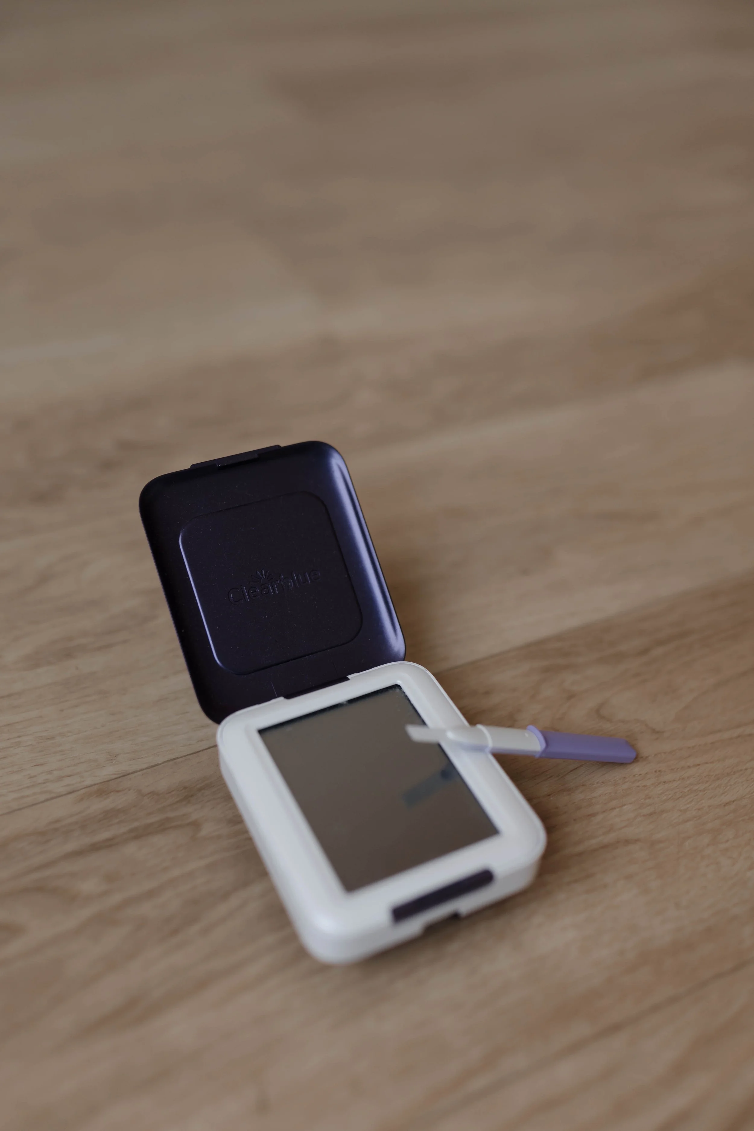 A small white glucometer with a test strip inserted is sitting on a wooden surface, with its black lid open.