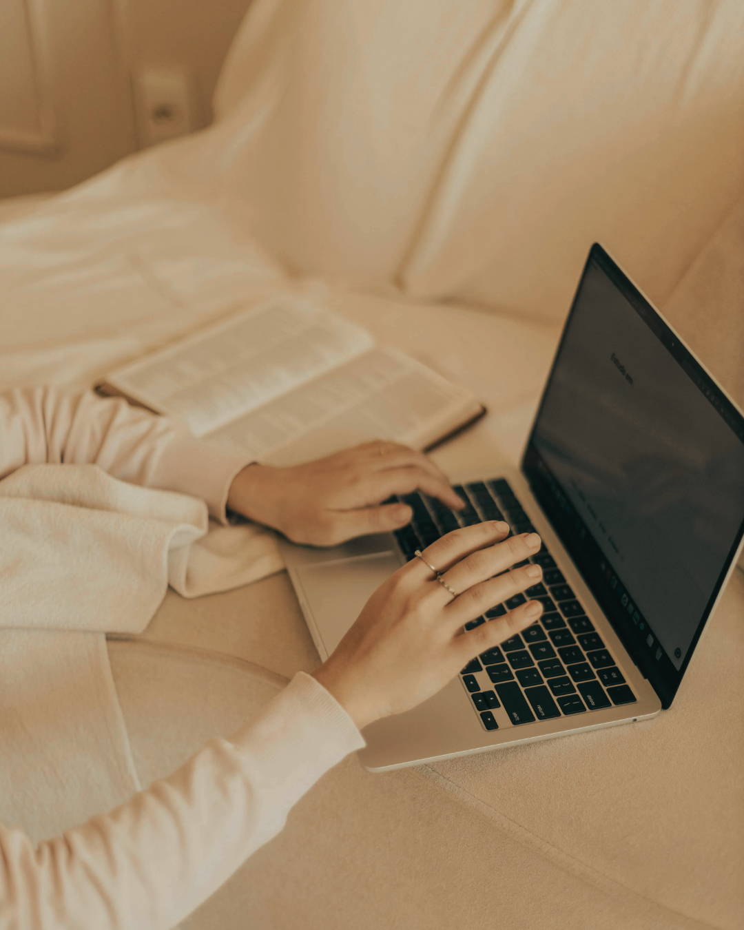 Person using a laptop computer while sitting on a bed, with an open book beside them.