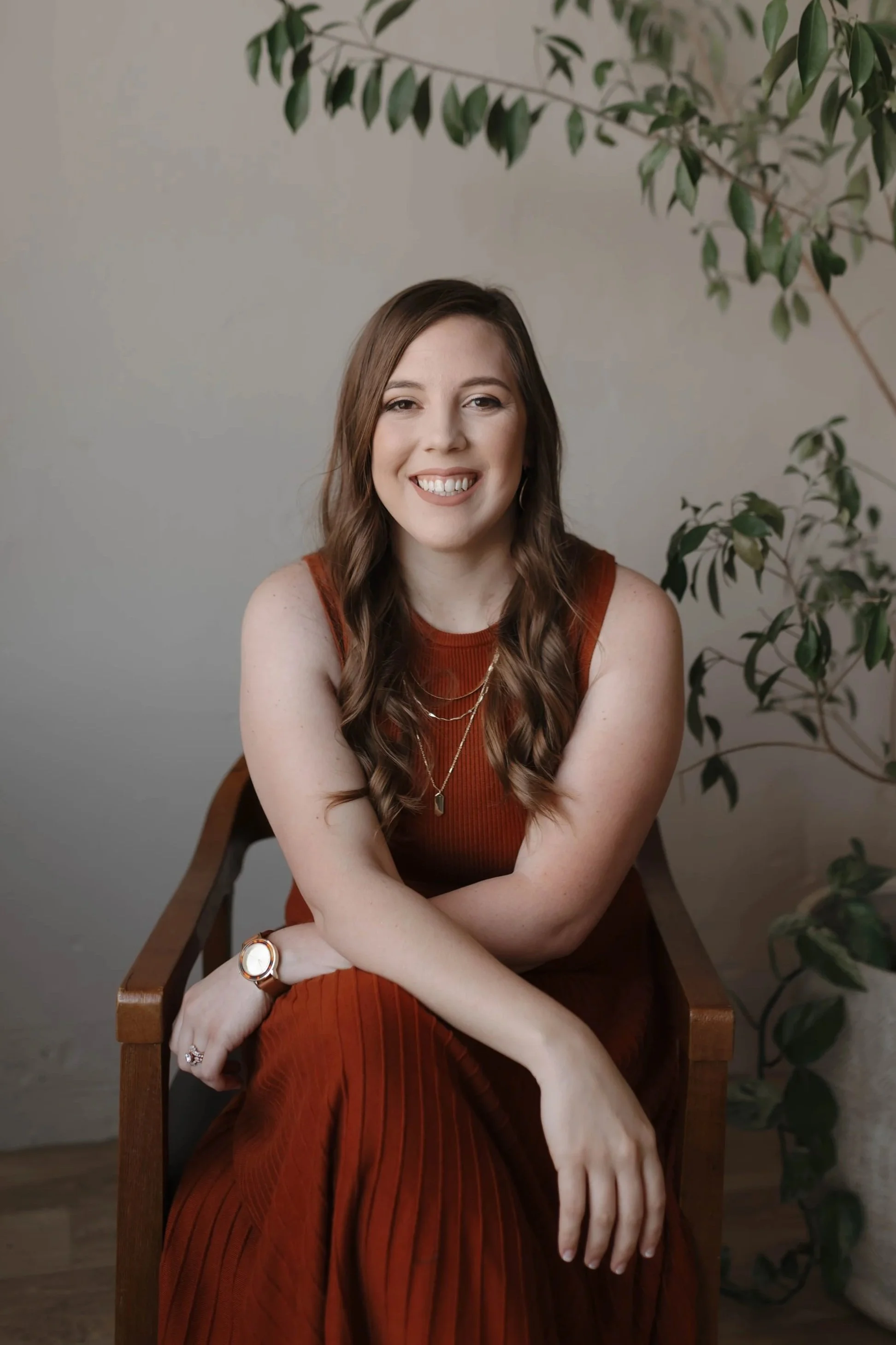 A woman with brown, wavy hair smiling, sitting on a wooden bench, wearing a rust-colored sleeveless dress, jewelry including a watch, rings, and layered necklaces, with green leafy plants in the background.