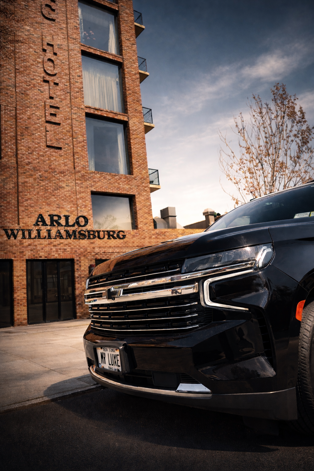 A black Honda car parked in front of a brick building with the sign 'ARLO WILLIAMSBURG'. The building has large windows and small balconies, with a tree on the right side and a cloudy sky above.
