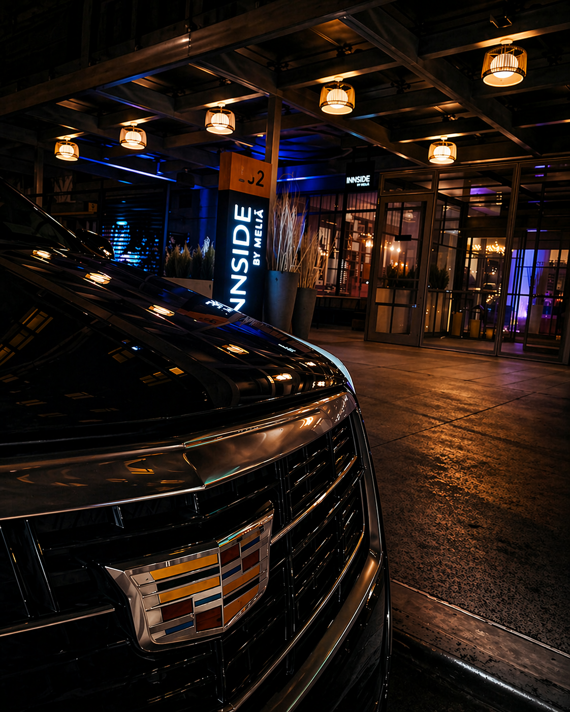 Close-up of a black Cadillac car parked outside a modern venue at night with illuminated signage that reads "INNSIDE BY MELIA" and a glass entrance door, with warm lighting overhead and ornamental plants near the entrance.