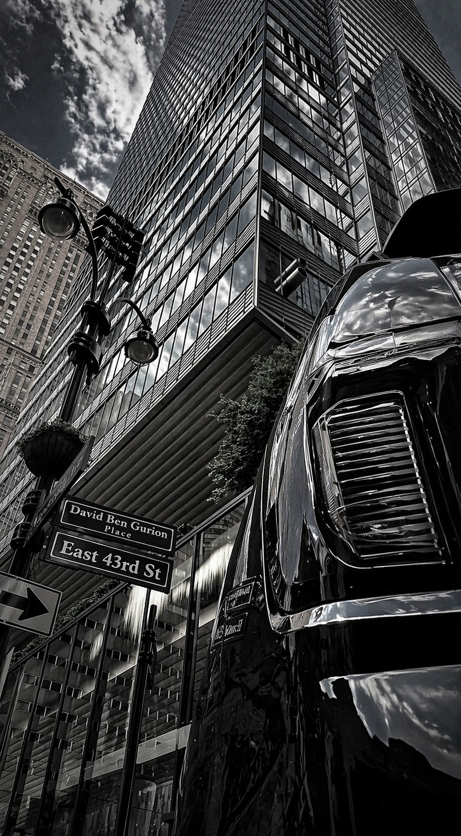 Black and white photo of a tall modern glass skyscraper, street signs, a streetlight, and part of a shiny vehicle in an urban city setting.