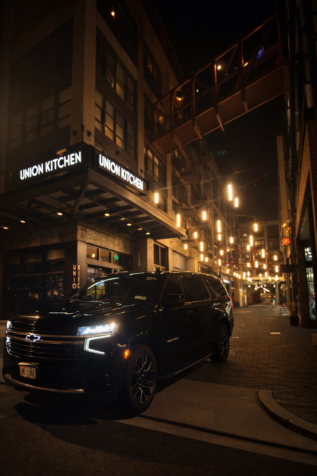 Night scene of a city street with a black Chevrolet SUV parked outside a restaurant named Union Kitchen, illuminated by warm hanging lights and signs.