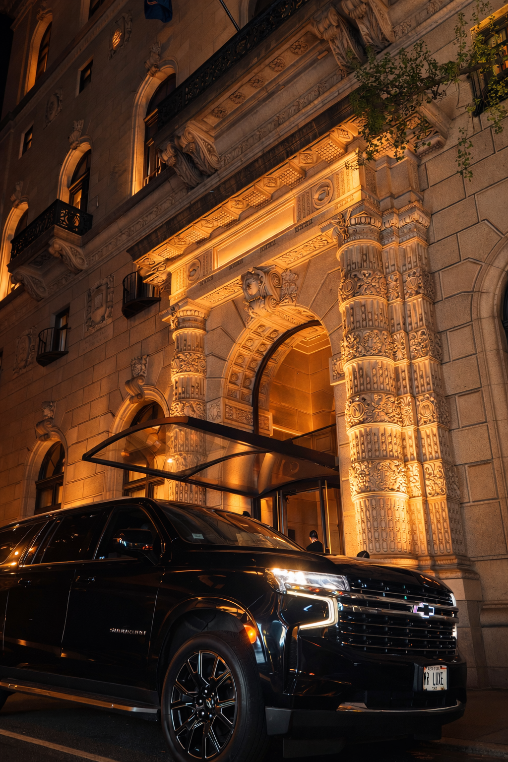 A black Chevrolet Suburban parked in front of an ornate, illuminated building entrance at night. The building features detailed stone carvings, columns, arches, and decorative balconies.