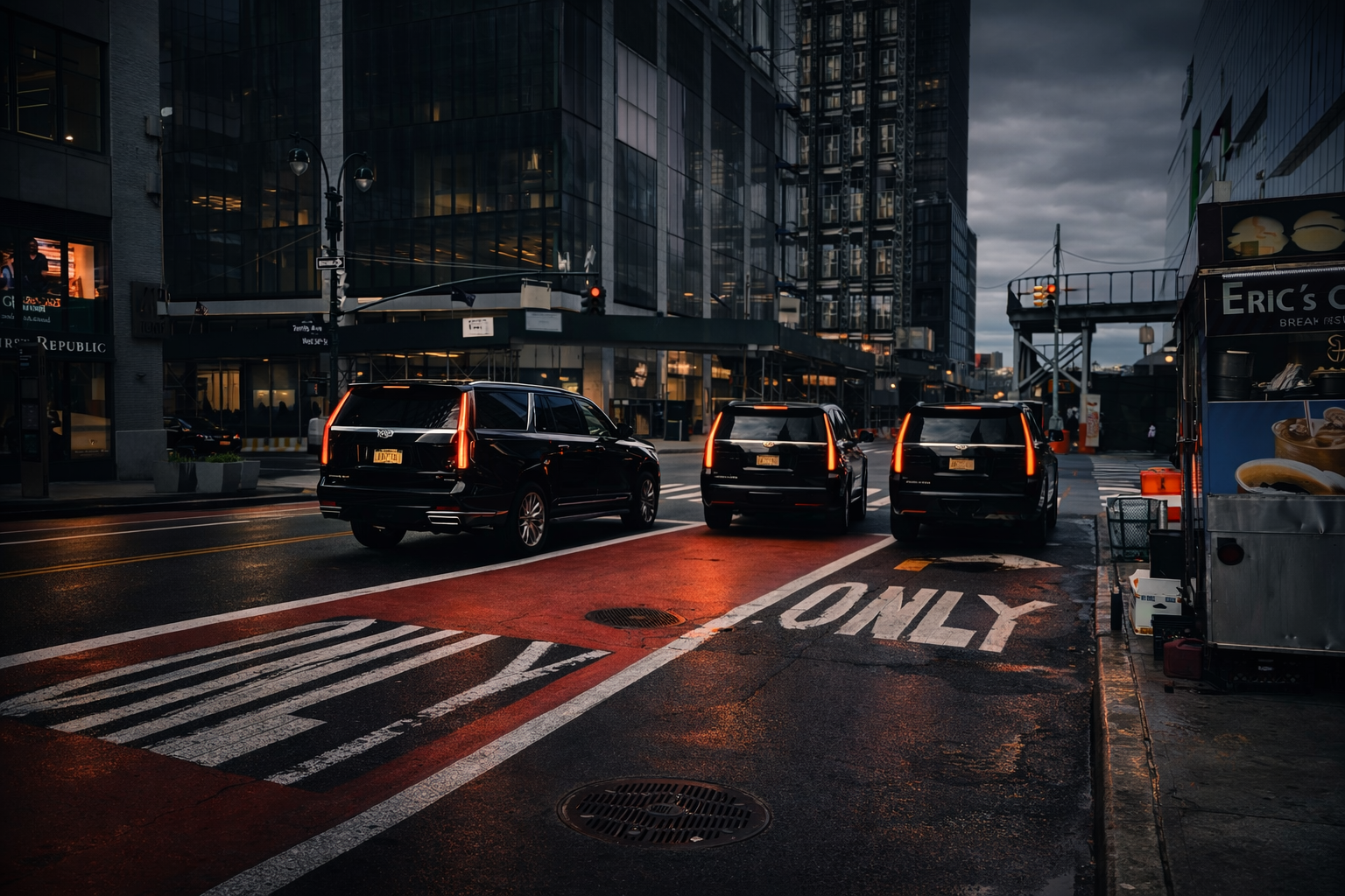 A city street scene at dusk with three black SUVs waiting at a red traffic light, surrounded by tall glass buildings and a food truck on the right side of the street