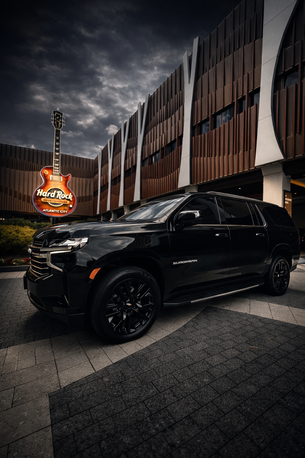 A black Chevrolet Suburban SUV parked in front of the Hard Rock Atlantic City building during dusk, with a sky filled with clouds in the background.
