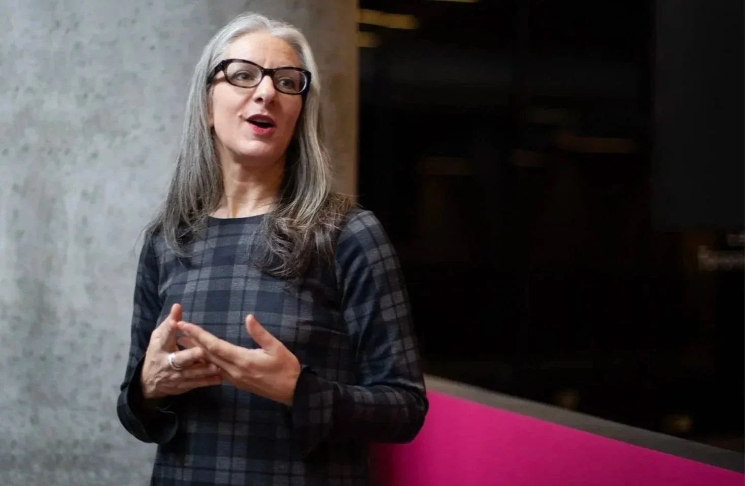 A woman with gray hair, wearing black glasses, and a dark plaid dress, speaking and gesturing with her hands, standing against a concrete wall and a dark background.