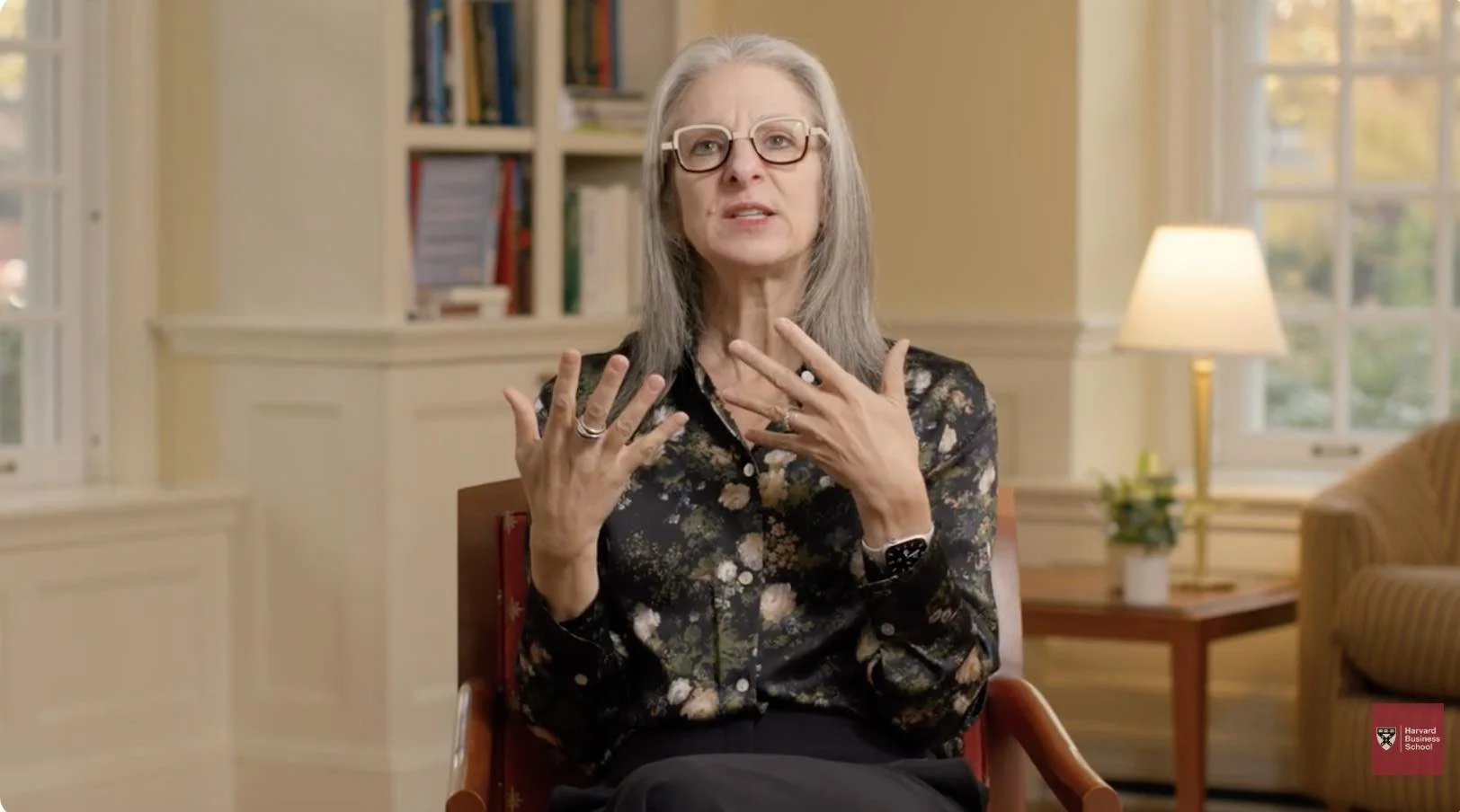 sarah kaplan with gray hair and glasses sitting in a library-style room gesturing with her hands as she speaks