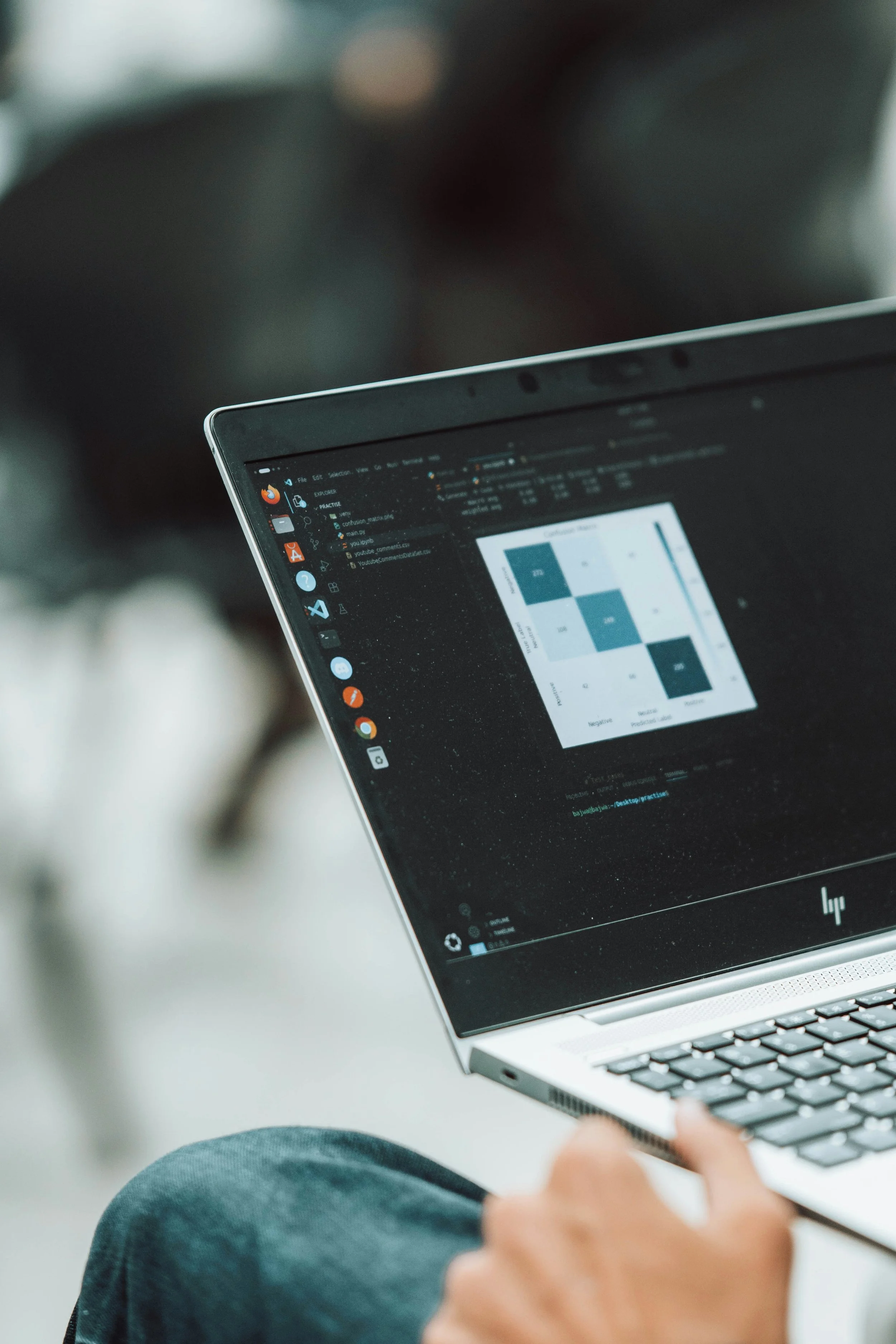 A person using a laptop displaying a dark-themed interface with various icons on the sidebar, including a color palette, settings, and a document. The person's hand is visible, and they are seated with their lap visible at the bottom of the image.