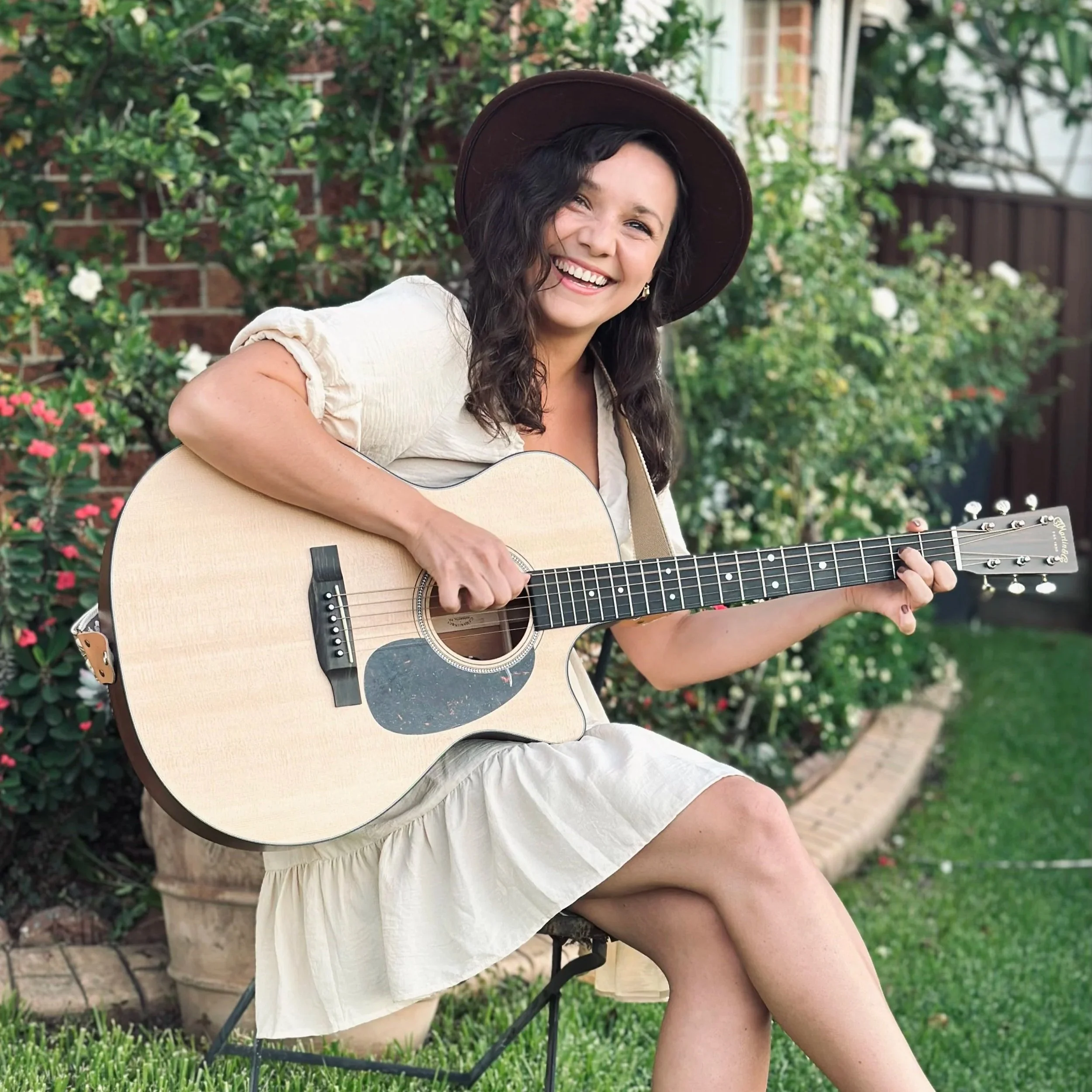 A woman with long dark hair wearing a wide-brimmed hat and a beige dress, playing an acoustic guitar outdoors in a garden with green plants and flowers in the background.