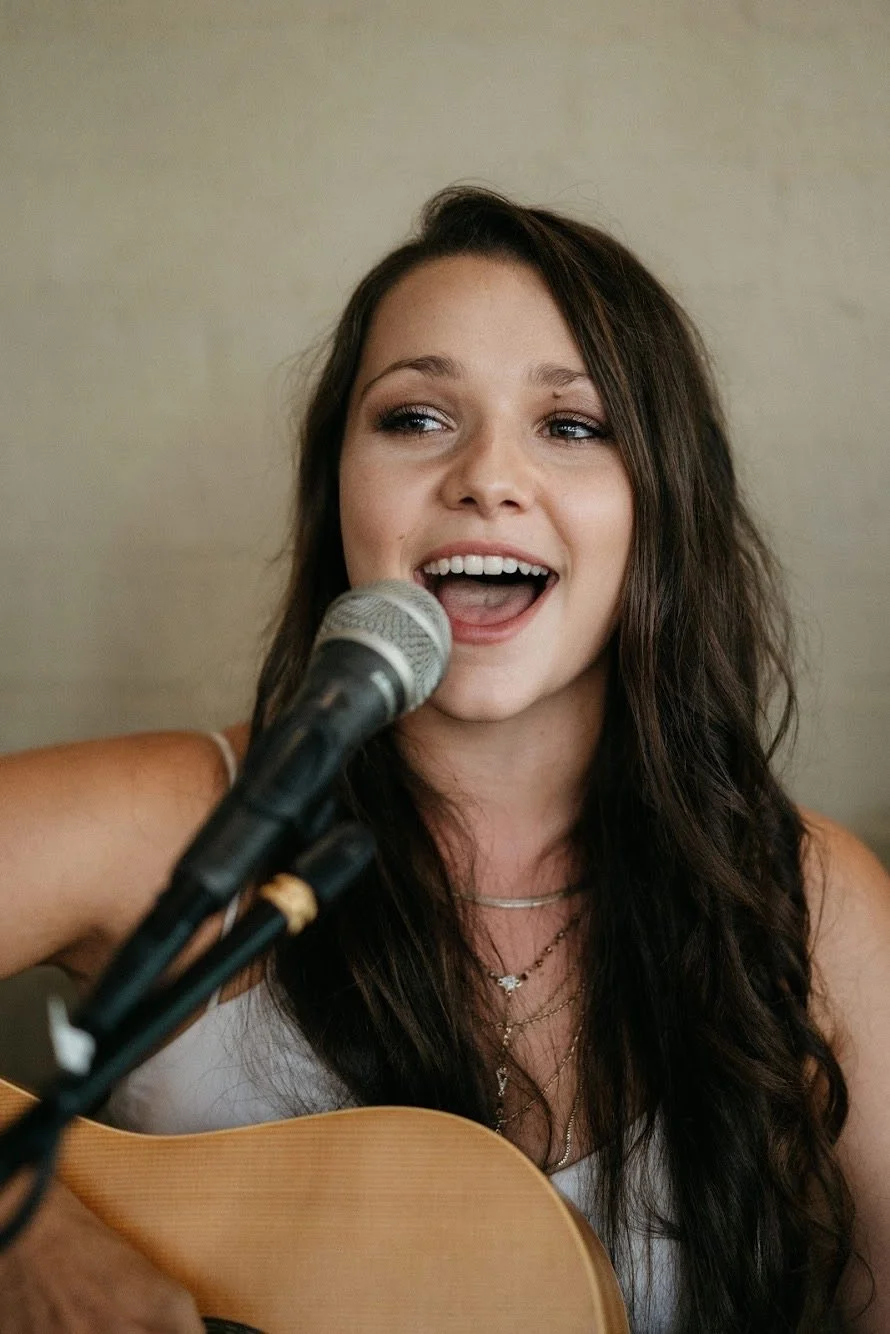 Young woman singing into a microphone while playing an acoustic guitar.
