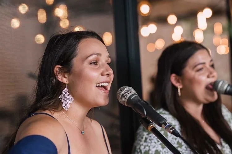 Two women singing into microphones during a performance with bokeh string lights in the background.