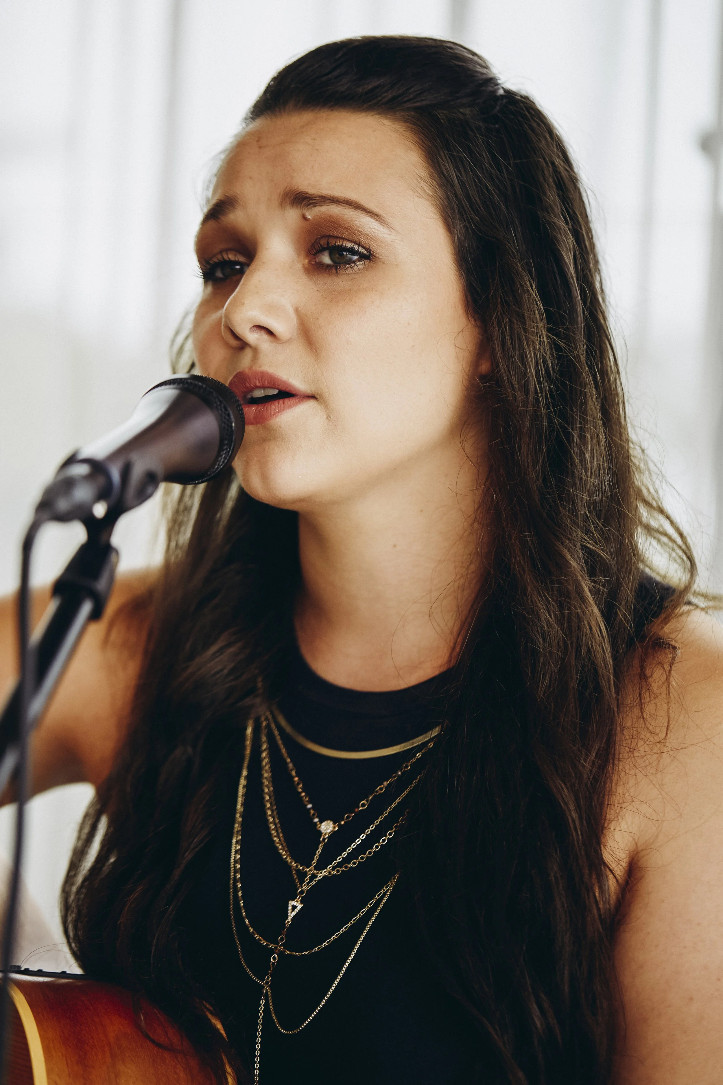 A woman singing into a microphone while strumming a guitar, with long wavy brown hair and wearing layered gold necklaces.