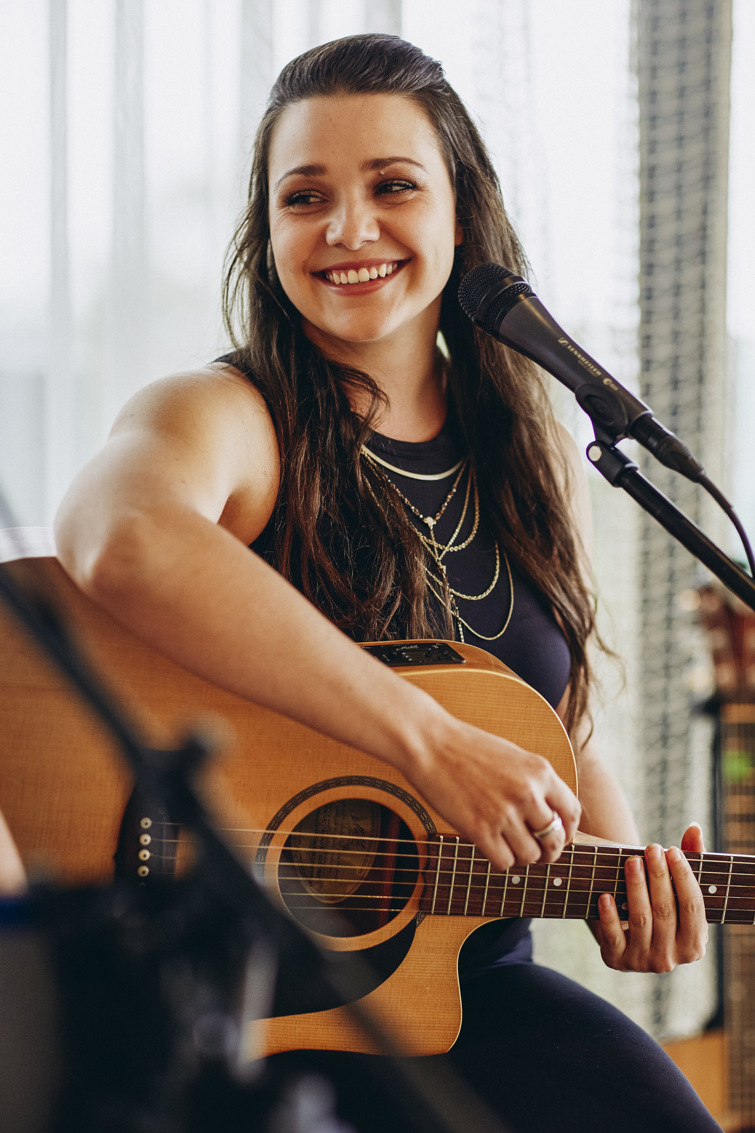 A woman with long dark hair smiling while playing an acoustic guitar and singing into a microphone.